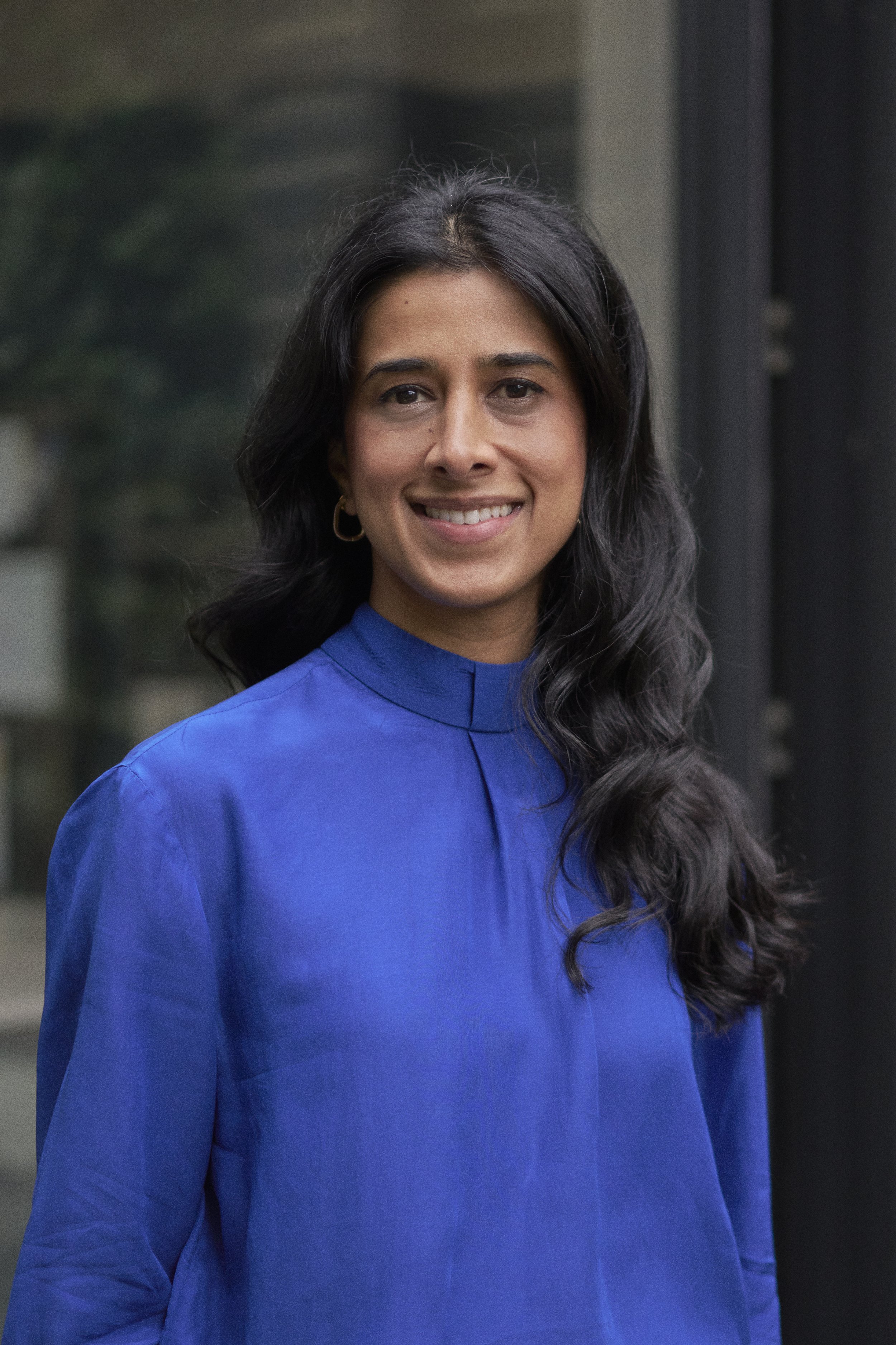 A woman with dark curly hair, wearing a royal blue top, smiling outdoors with blurred background.