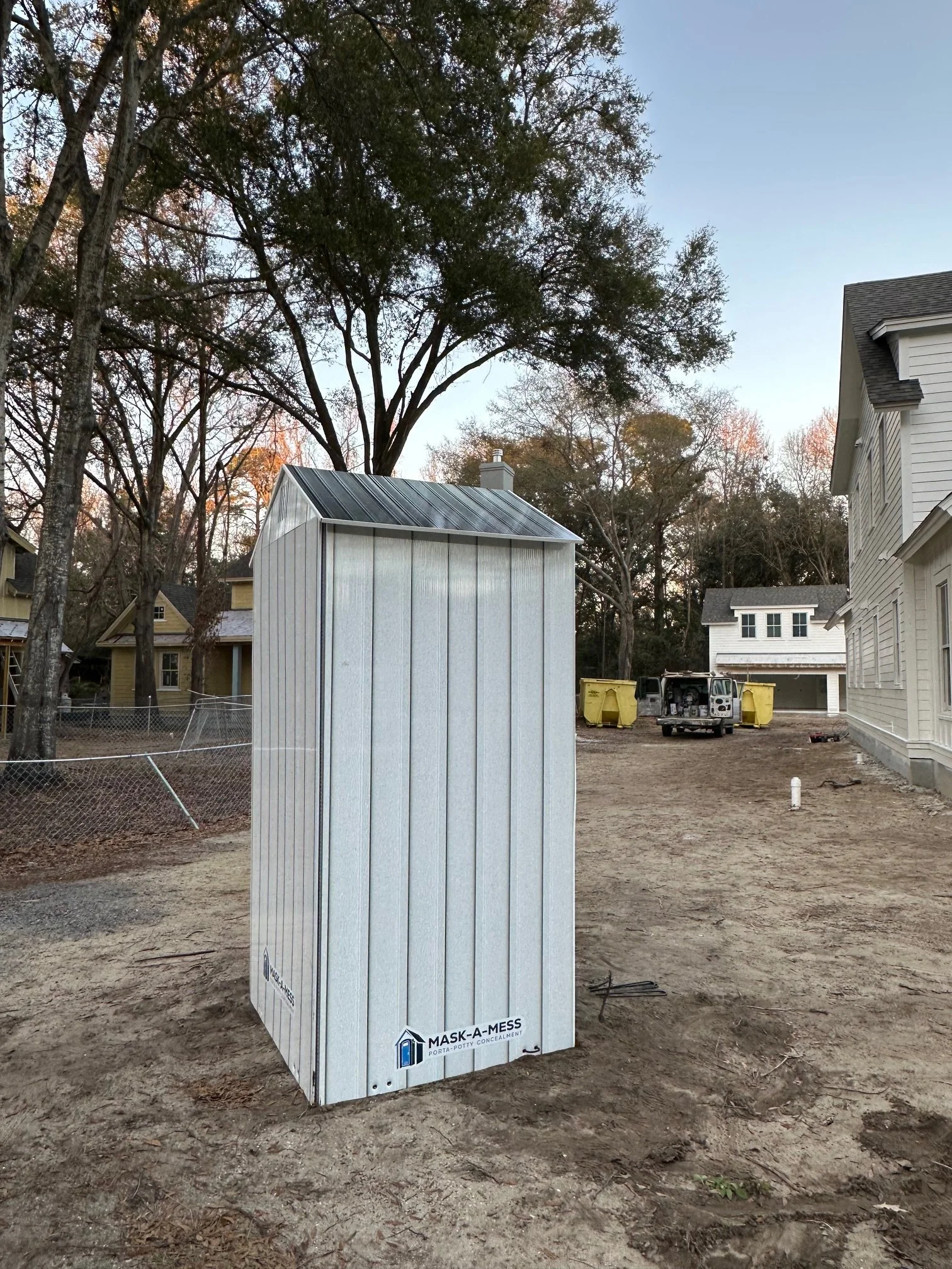 A portable toilet labeled 'Mask-A-Mess' on a construction site with dirt ground, surrounded by trees and residential buildings in the background.