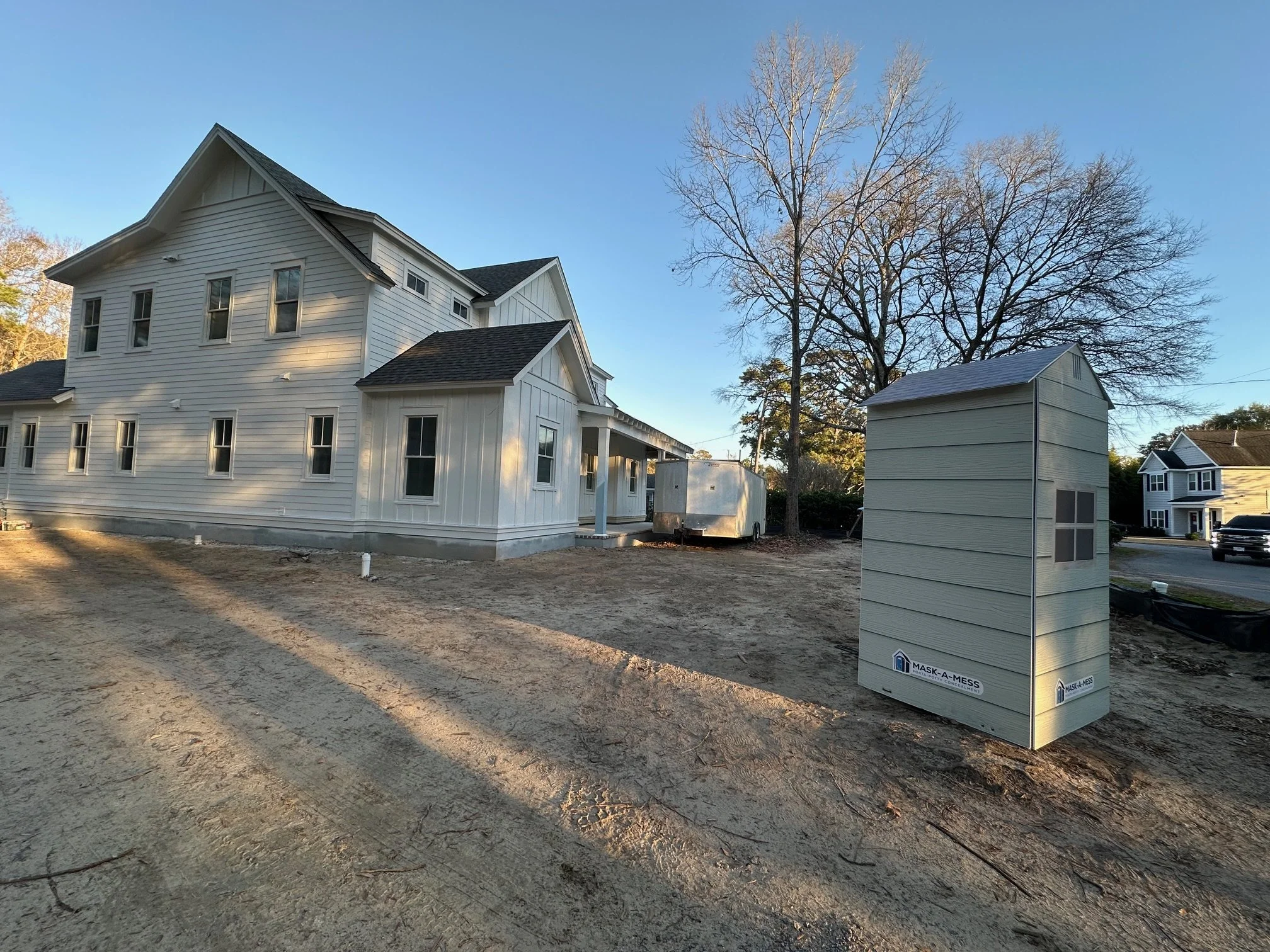 A new white house under construction with a small white storage shed in front and a large tree behind the house. There is a trailer next to the house and the ground is dirt.