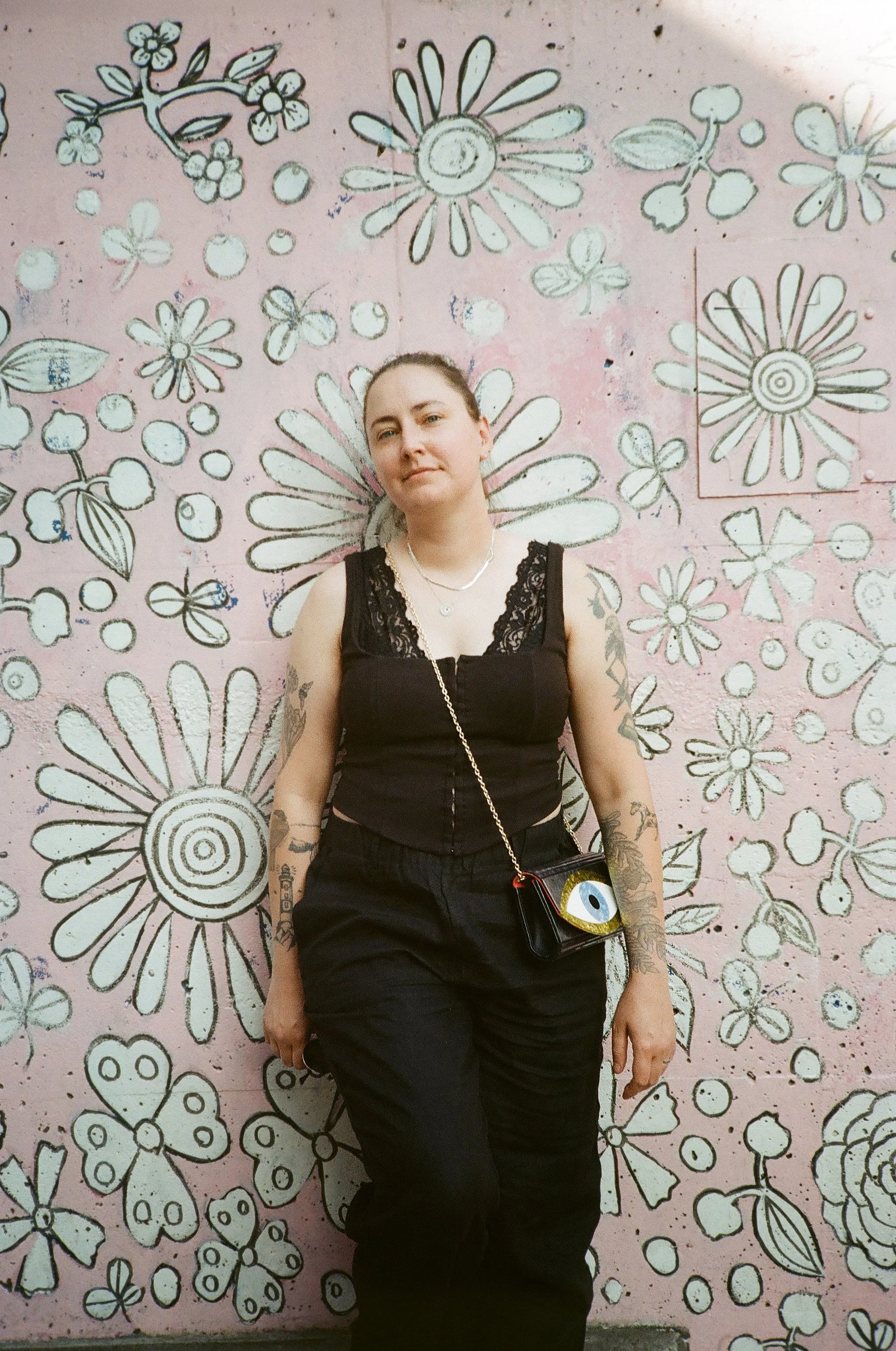 Photo of Sara wearing an all black outfit leaning against a pale pink wall painted with black and white flowers
