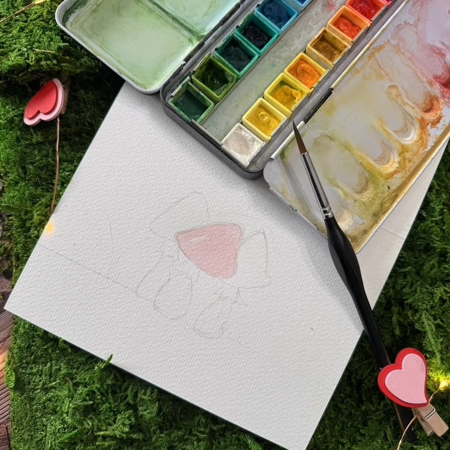 Watercolors, a paintbrush, and a blank paper with a faint outline of a heart-shaped butterfly. The painting supplies are on a green surface with small decorative pink and red hearts.