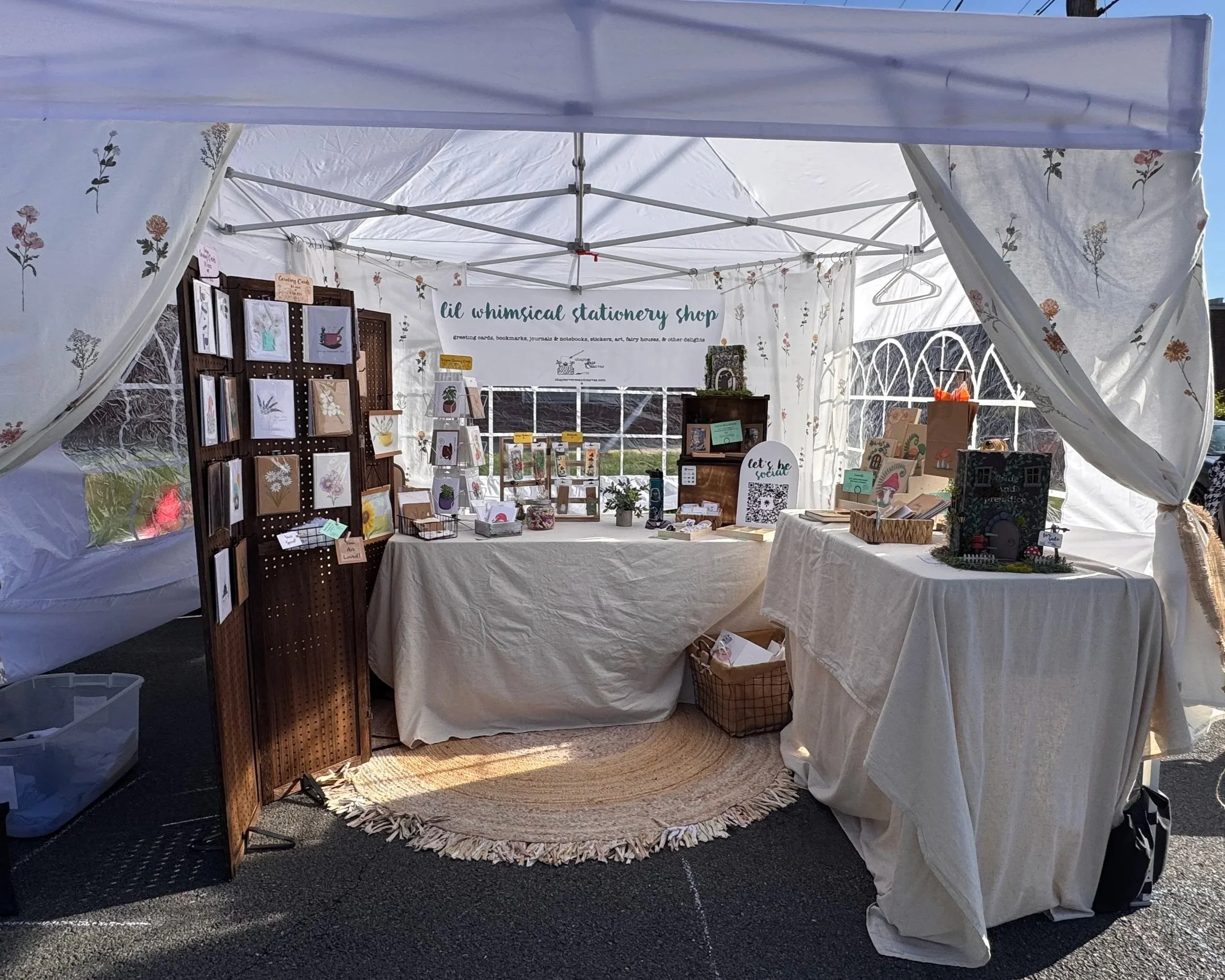 Booth at an outdoor craft fair featuring stationery and artwork, decorated with floral-themed displays, small plants, and a woven rug.