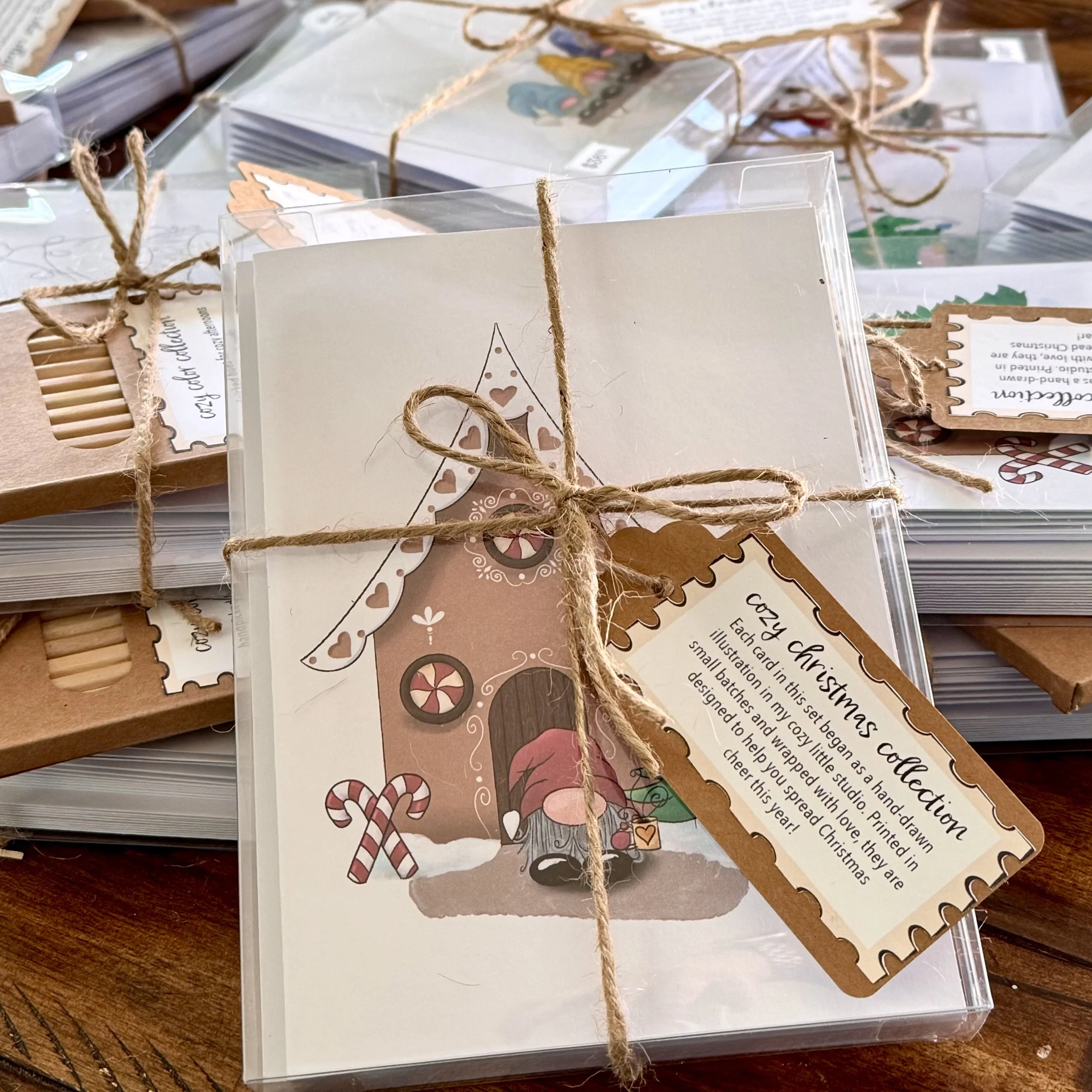 A wrapped Christmas card featuring a cute illustration of a gingerbread house, decorated with candy canes, lollipops, and a gnome wearing a Santa hat, tied with twine on a wooden surface.