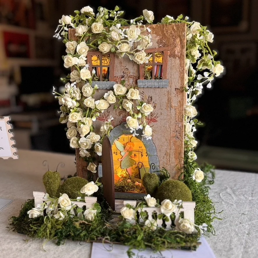 A decorative miniature scene featuring a weathered pinkish-brown building with small windows, surrounded by white roses and greenery. There is a small fairy figure illuminated inside a circular opening with a window, alongside moss-covered rocks and two tiny white fences.