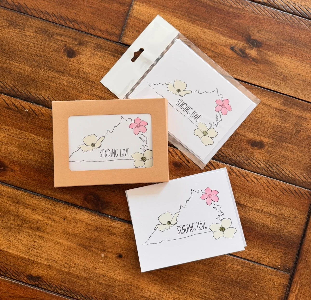Set of greeting cards with a floral outline of Hawaii and the text "Sending Love." One card is in a peach-colored box, and others are outside the box on a wooden surface.