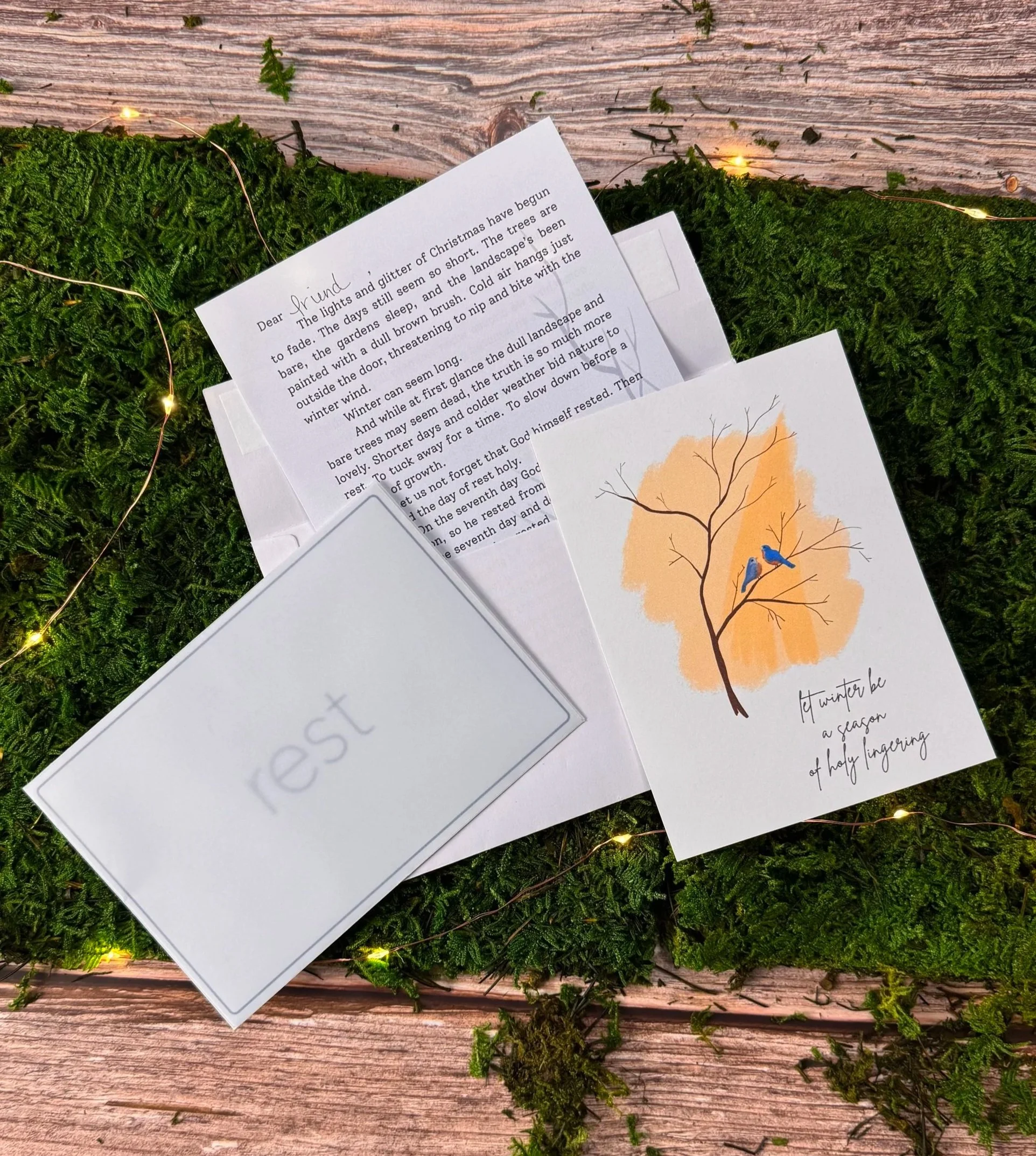 A Christmas scene with a moss-covered surface decorated with tiny string lights, an open letter, a white card with the word 'rest' faintly written on it, and a holiday card featuring an illustration of a tree with two bluebirds and the message 'If winter be a season of holy longing.'