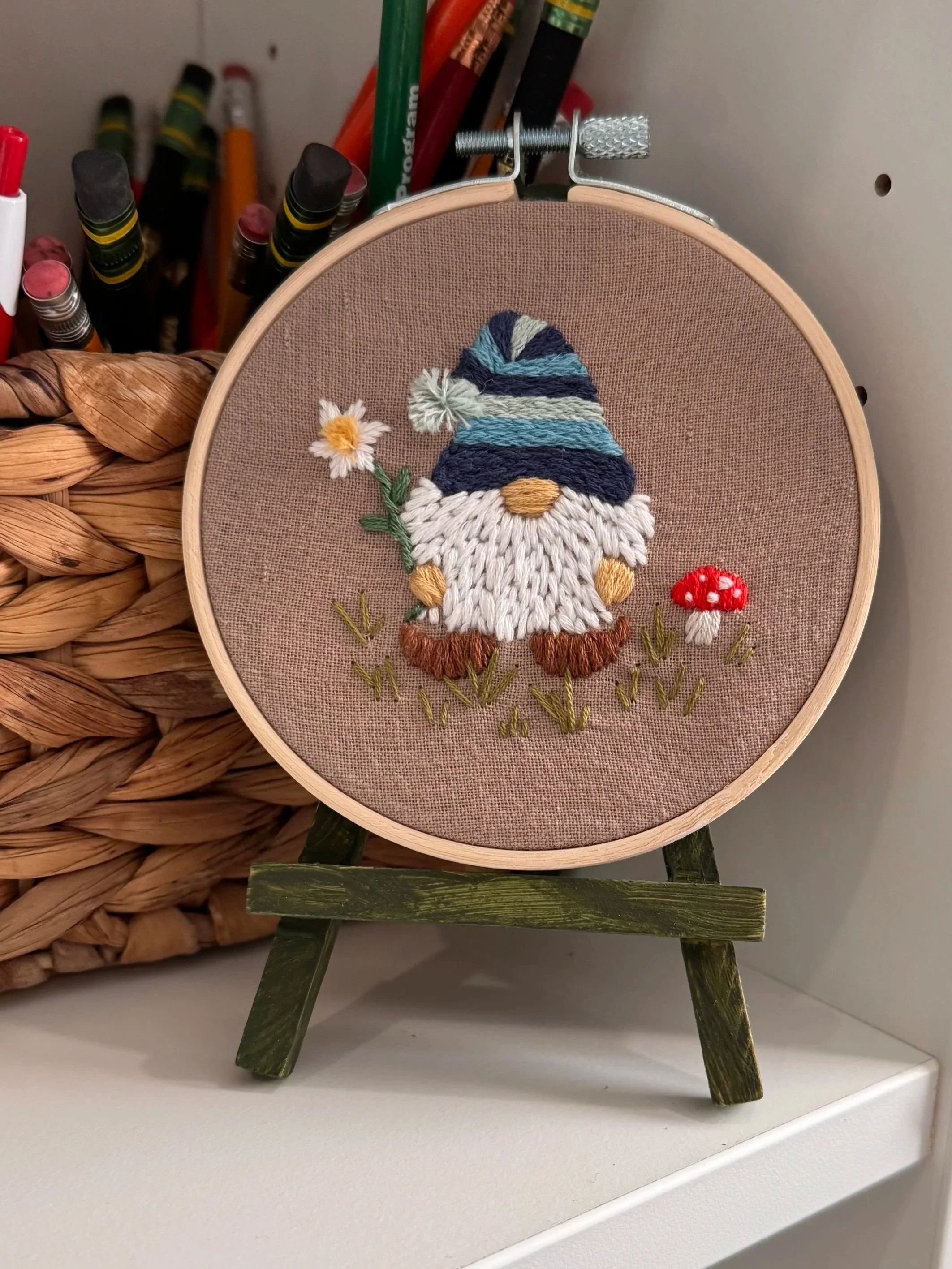 Embroidery of a garden gnome holding a flower, wearing a blue-striped hat, with mushrooms nearby, on fabric stretched in a hoop.