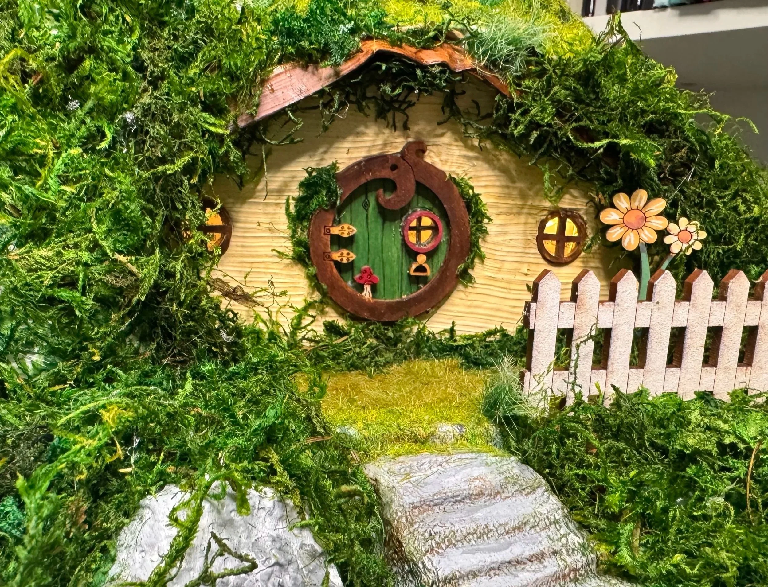 A miniature fairy house with a green door and round windows, surrounded by lush green moss and plants, and decorated with a wooden fence and flower cutouts.