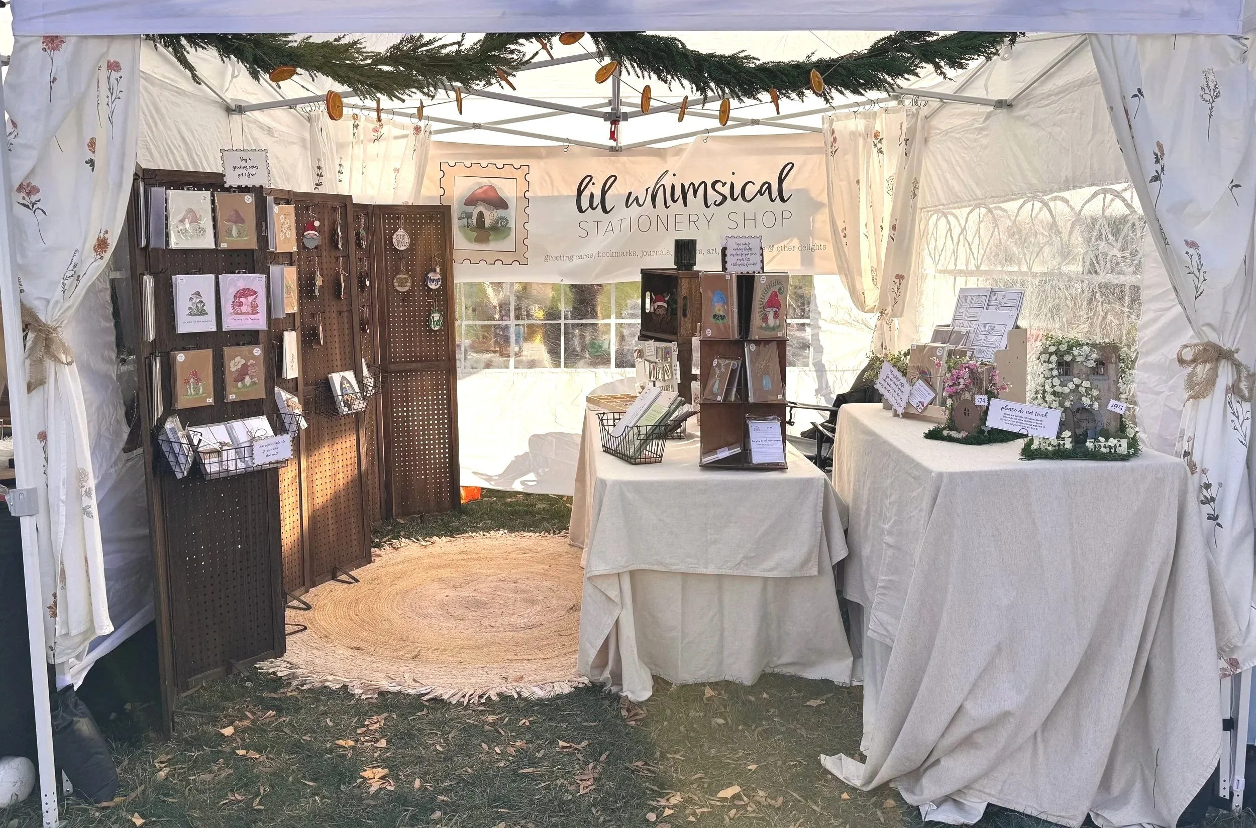 Inside a craft tent at an outdoor market, a stationeri shop called 'Lil Whimsical Stationery Shop' displays greeting cards, bookmarks, journals, and art items on racks and tables, with a white banner overhead and decorated with greenery and flowers.