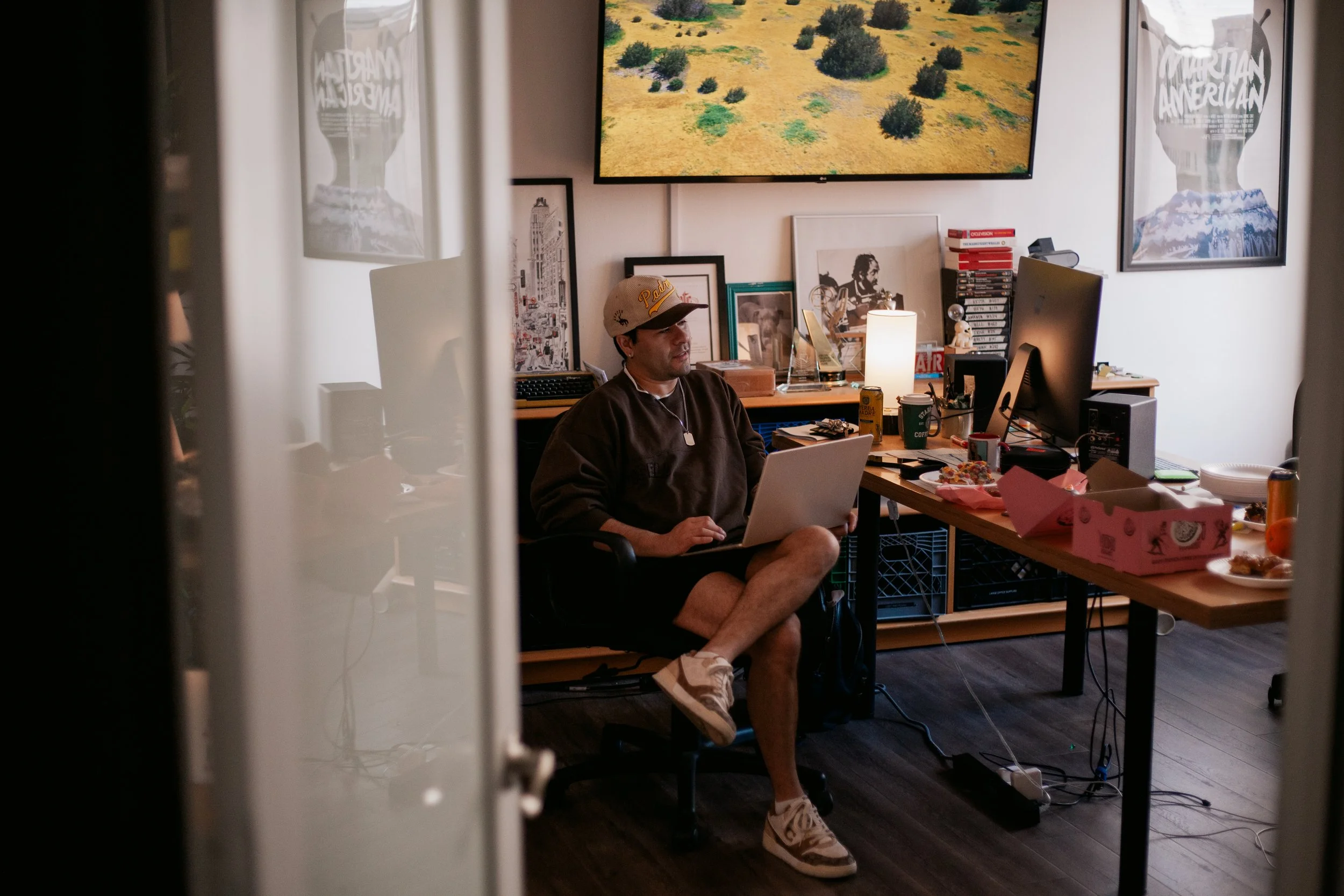 A man sitting at a desk in a room with wall art, a large TV screen displaying a landscape, and cluttered items, viewed through a slightly open door or curtain.