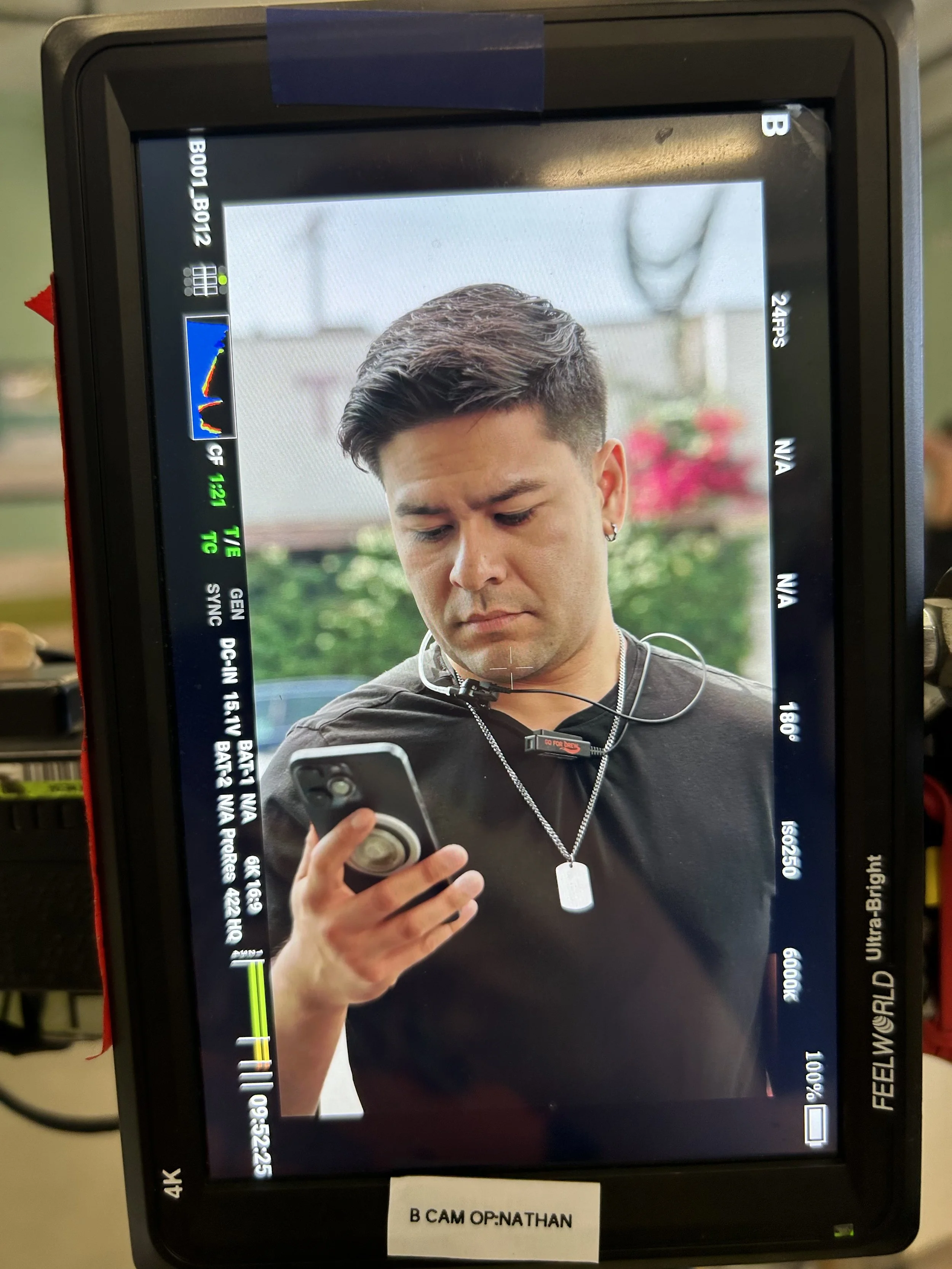 A monitor displaying a photo of a young man with styled hair and earrings, looking at his phone. The monitor has various settings and info on the edges, with a label below reading 'B CAM OP:NATHAN'.