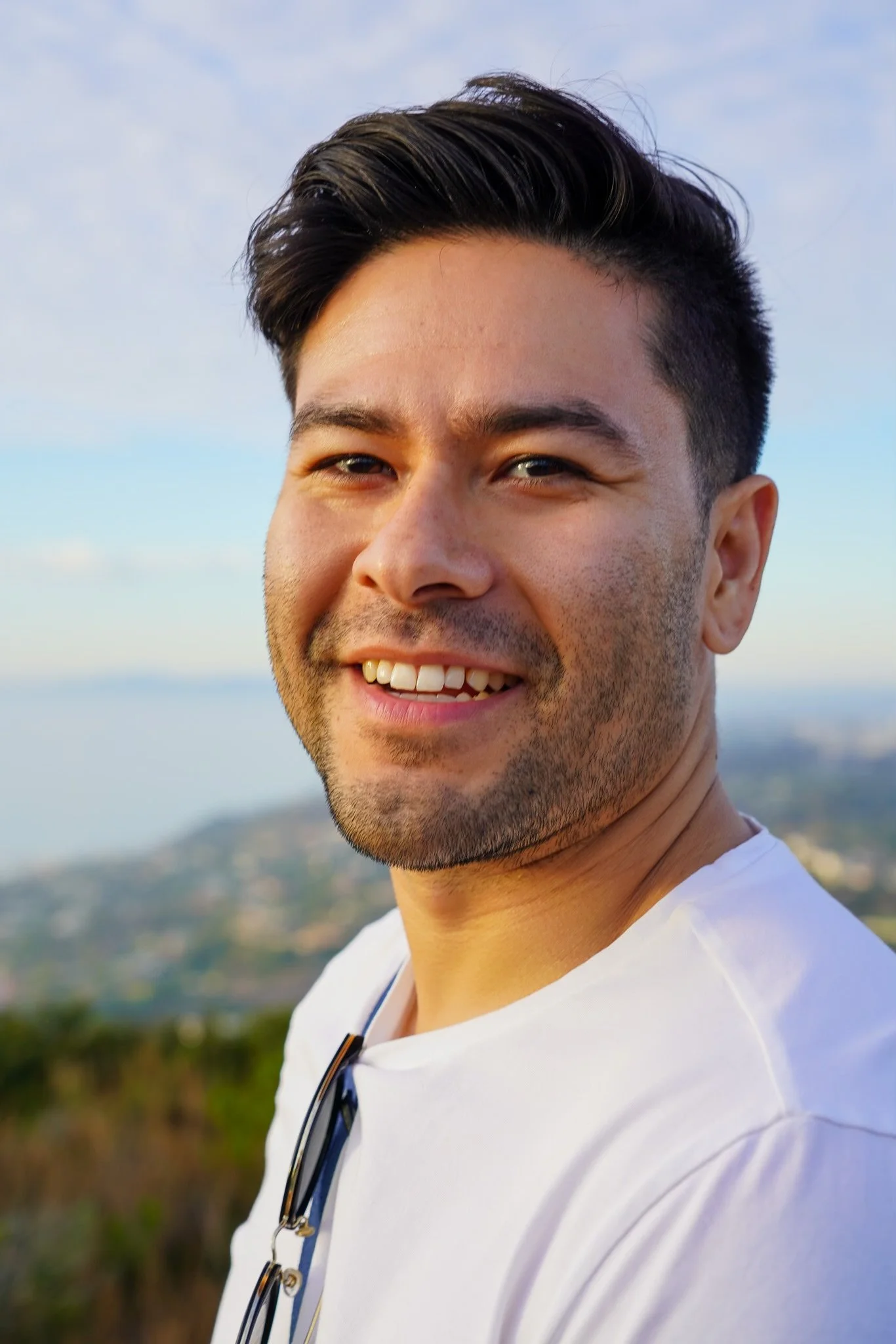 Close-up of a young man smiling outdoors during daytime, wearing a white shirt with sunglasses hanging from the collar, with a blurred landscape and sky in the background.