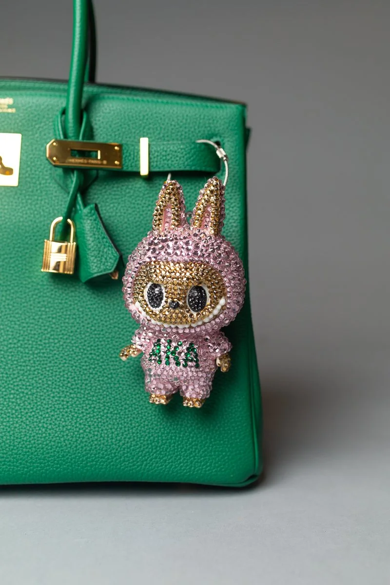 A pink rhinestone-encrusted keychain in the shape of a bunny with large black eyes, wearing a pink outfit with green letters that spell "AIKA". It is attached to a green handbag.