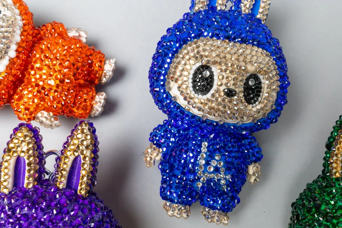 A collection of colorful, beaded animal keychains decorated with rhinestones, including a blue sloth in the center, an orange dog, a purple bunny, and a green animal, arranged on a white background.