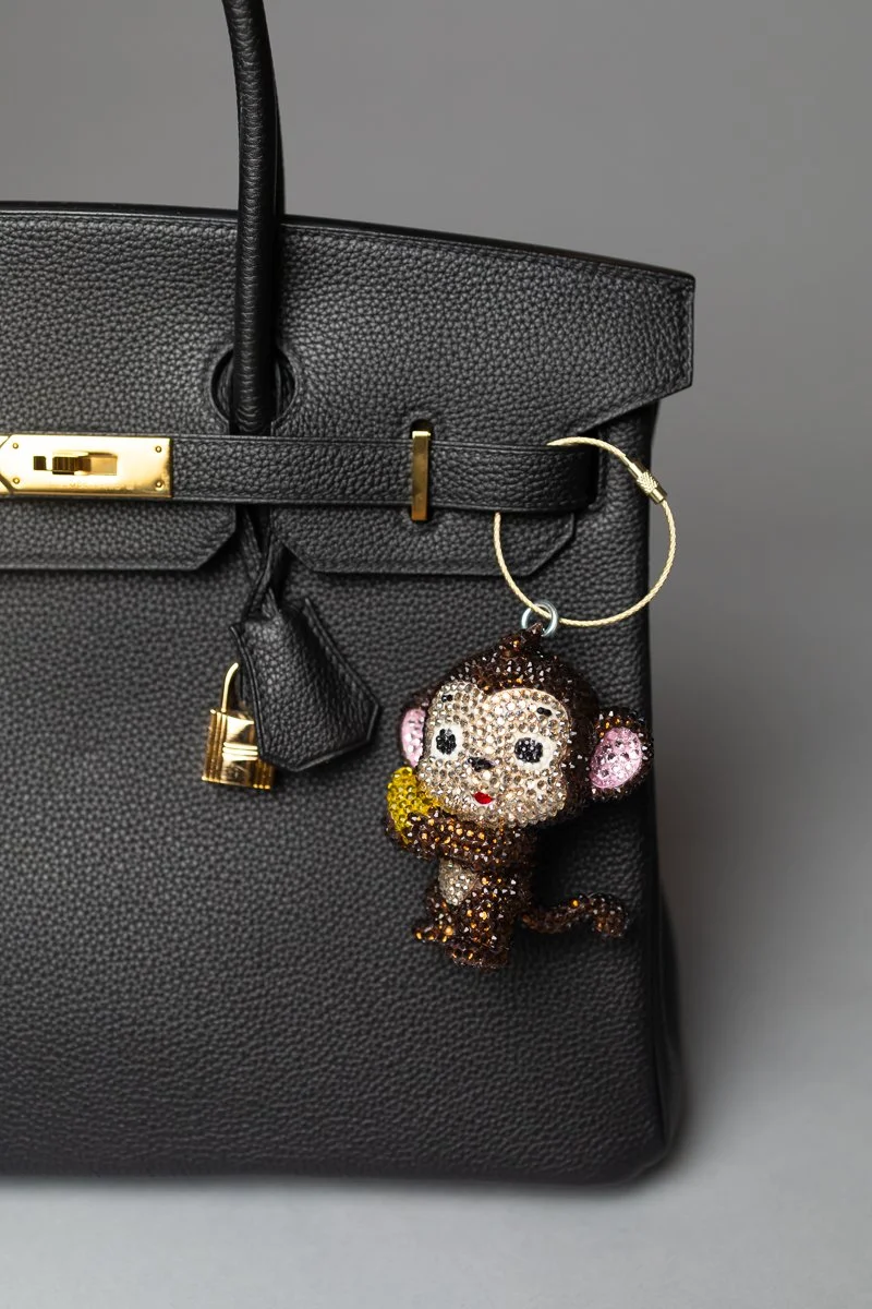 Black designer handbag with gold hardware and a monkey keychain adorned with rhinestones hanging from the handle.