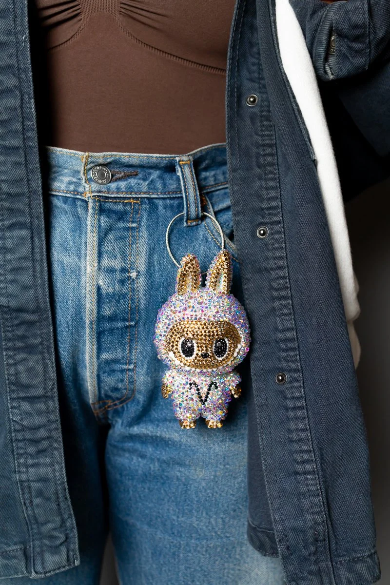 A close-up of a person wearing blue jeans and a brown top, with a colorful, rhinestone-encrusted bunny keychain hanging from the belt loop of the jeans.