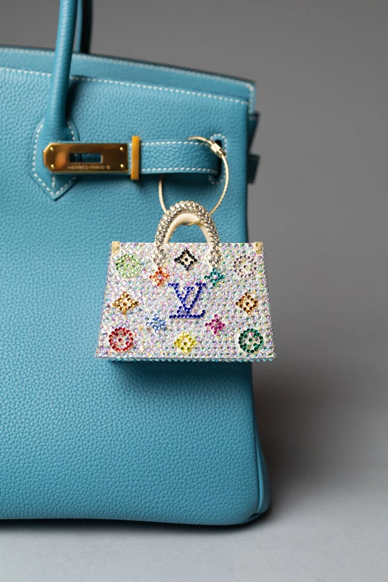 A blue handbag with a small, colorful, rhinestone-encrusted purse shaped like a handbag hanging from it.