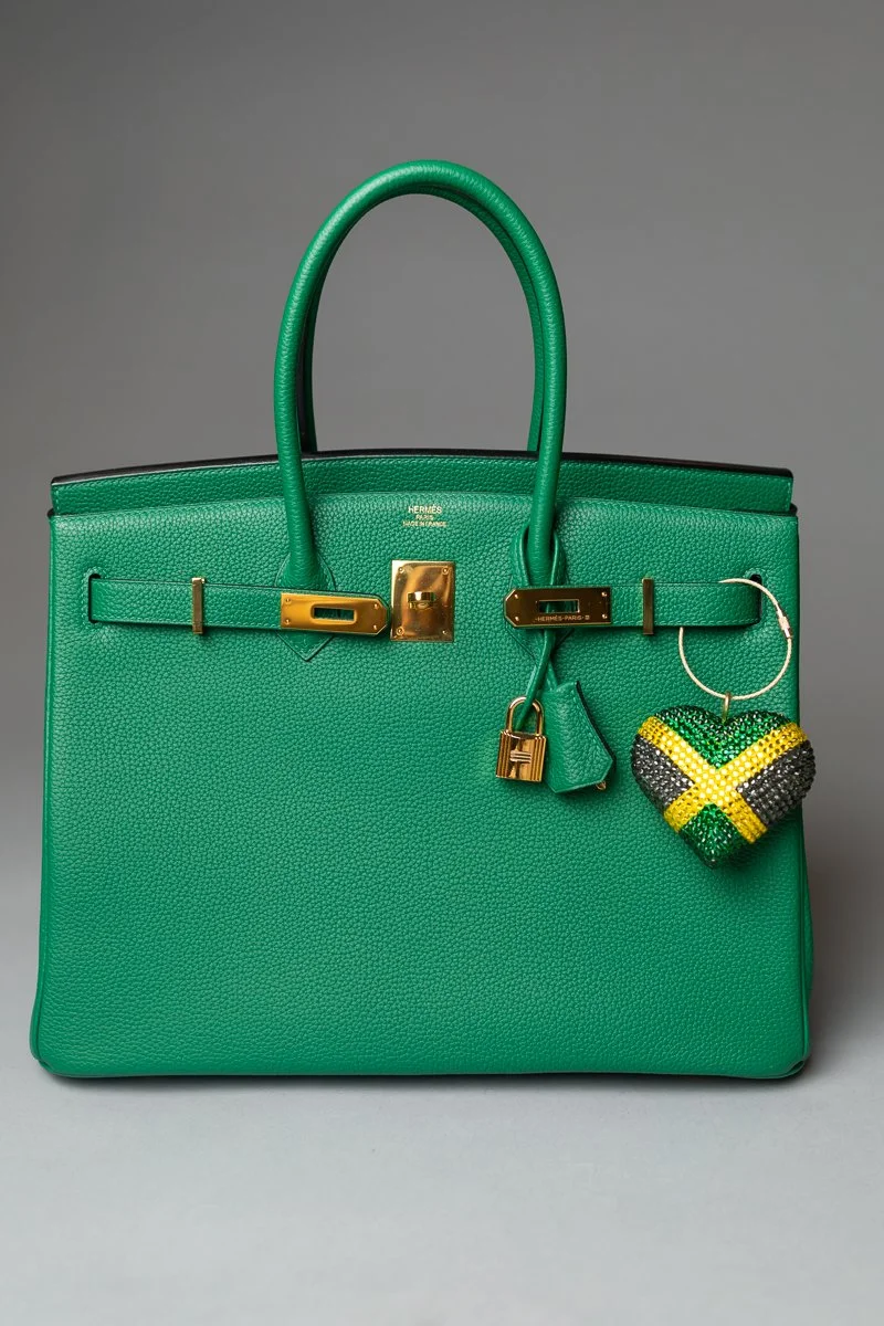 A green Hermès leather handbag with gold hardware, accompanied by a beaded heart-shaped keychain in green, yellow, black, and white, against a gray background.