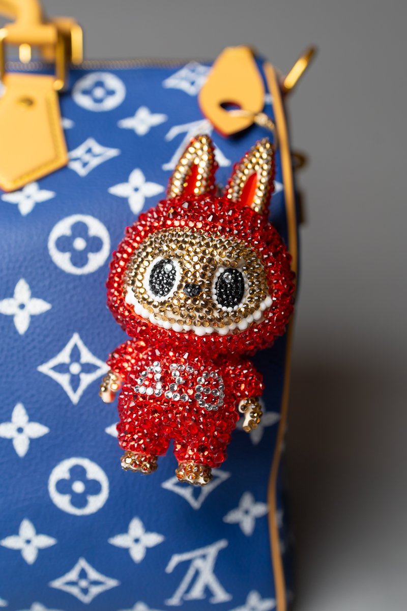 A small, red, bejeweled plush toy in the shape of a bunny with large black eyes, a tiny nose, and wearing a red jumpsuit with white accents. The plush is attached to a designer blue and white patterned handbag with gold hardware.