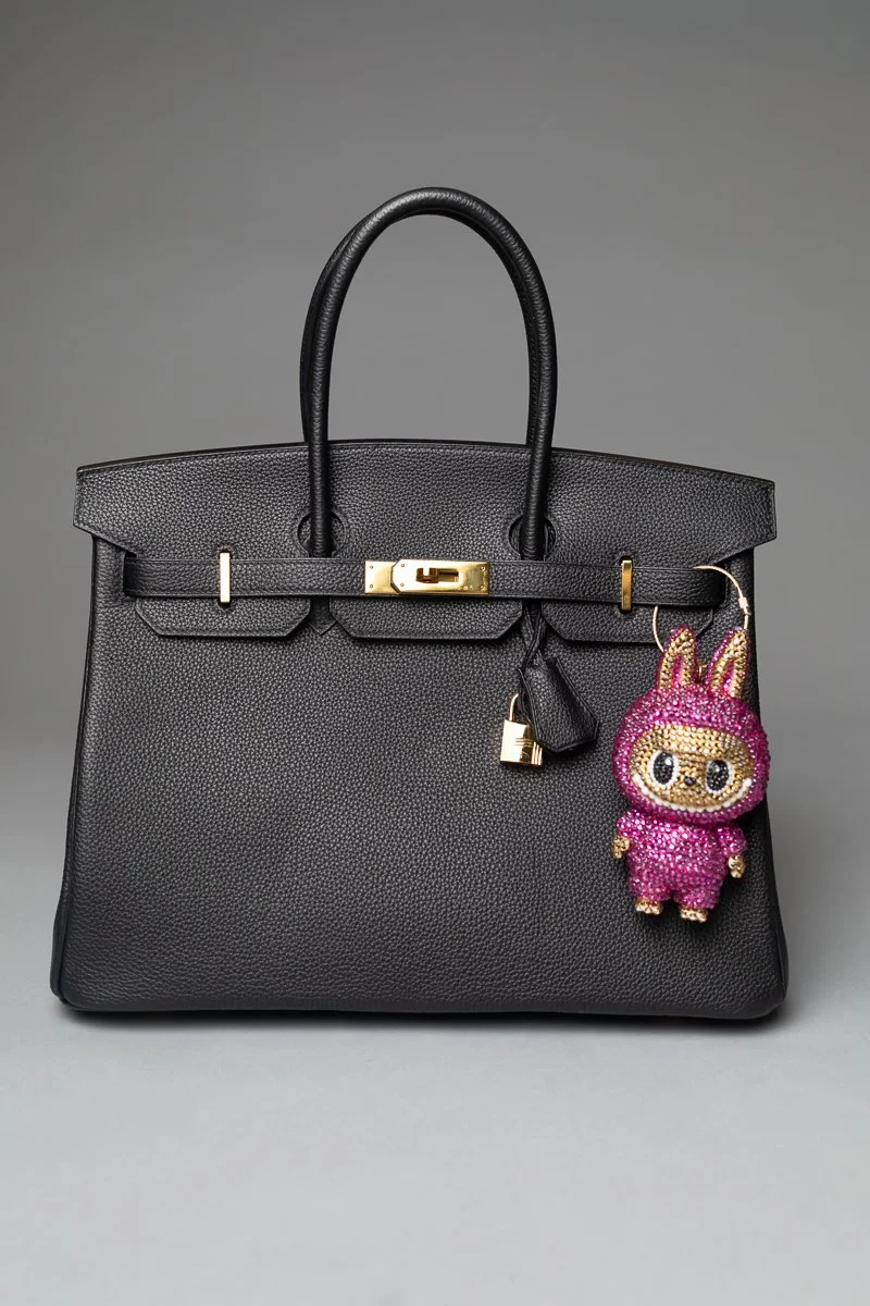 A black leather handbag with gold hardware and a pink, beaded plush keychain character hanging from it, set against a gray background.