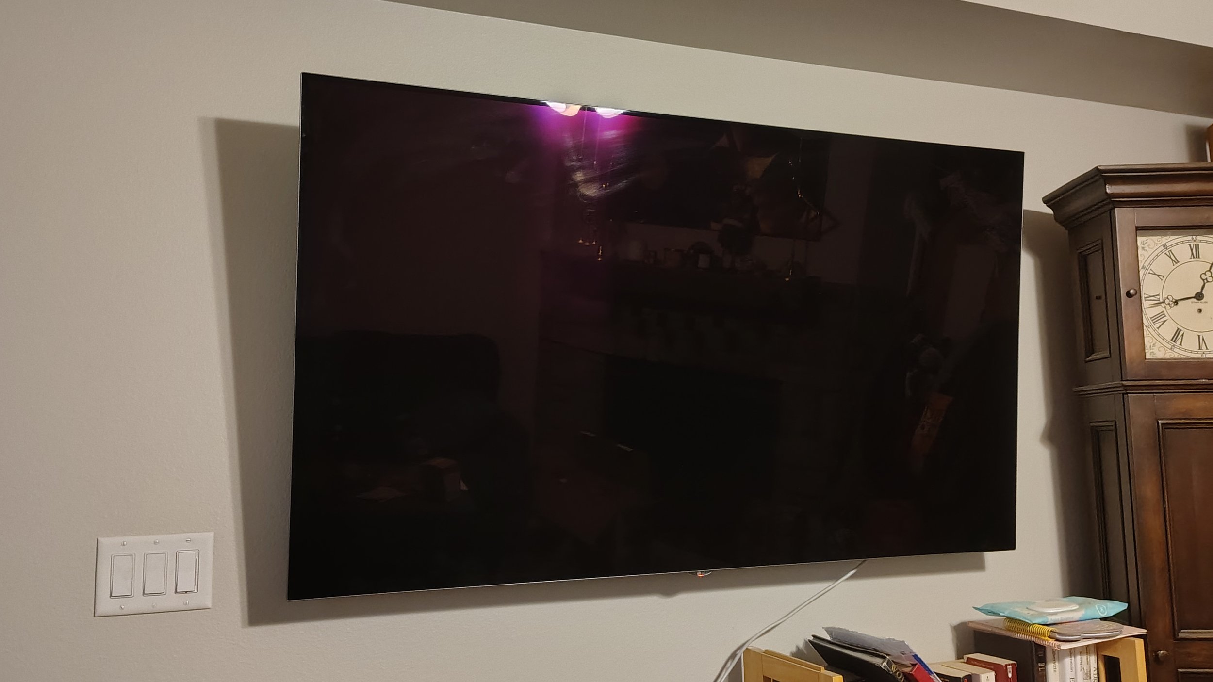 A large, wall-mounted flat-screen television turned off in a living room. There is a reflection of the room visible on the screen. To the right of the TV is an antique wooden clock cabinet, and below it are some books, tissues, and papers.