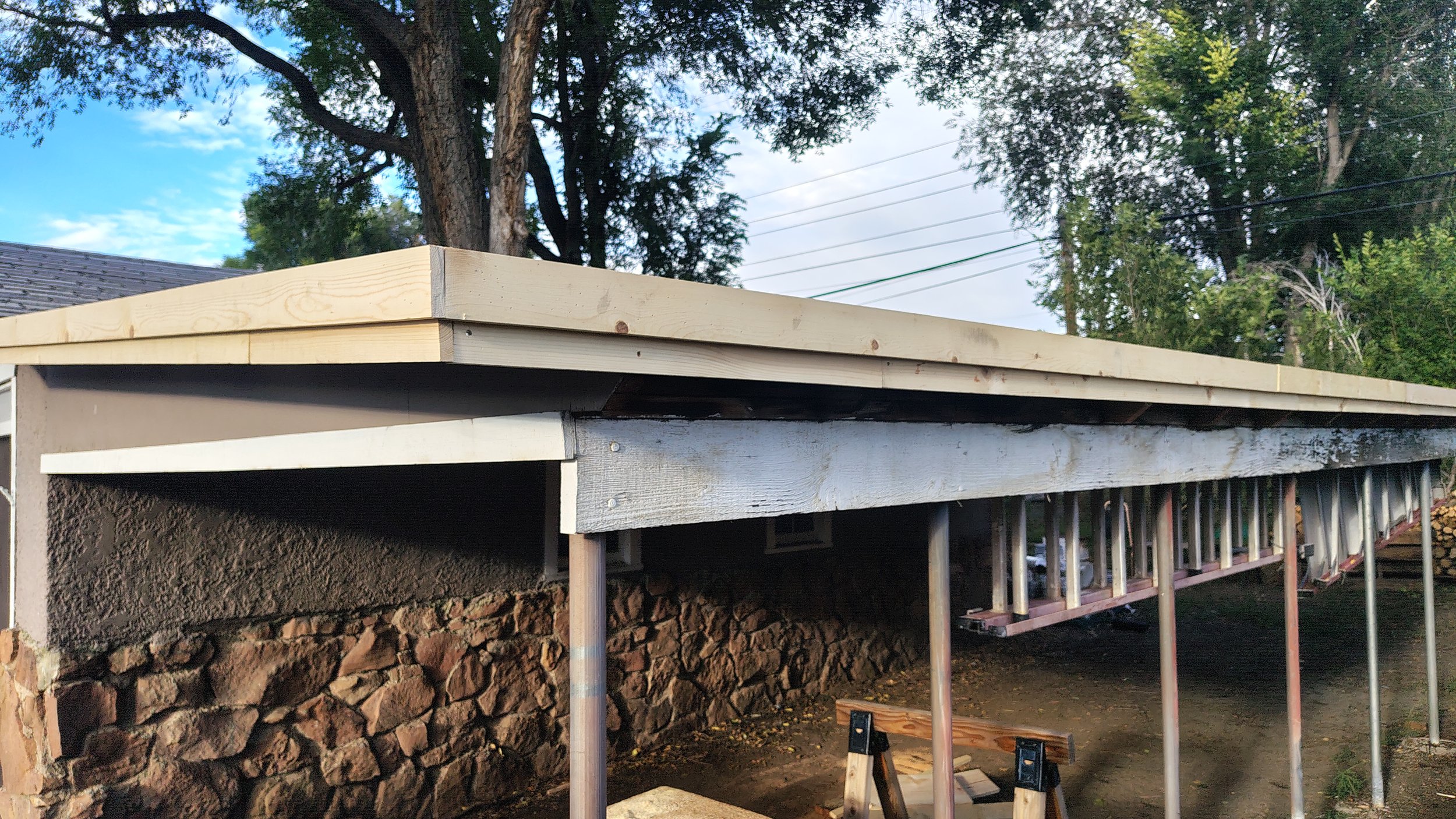 A wooden deck under construction with a new light-colored wooden top, supported by metal posts. The deck is built next to a stone foundation and a house, with trees and a partly cloudy sky in the background.