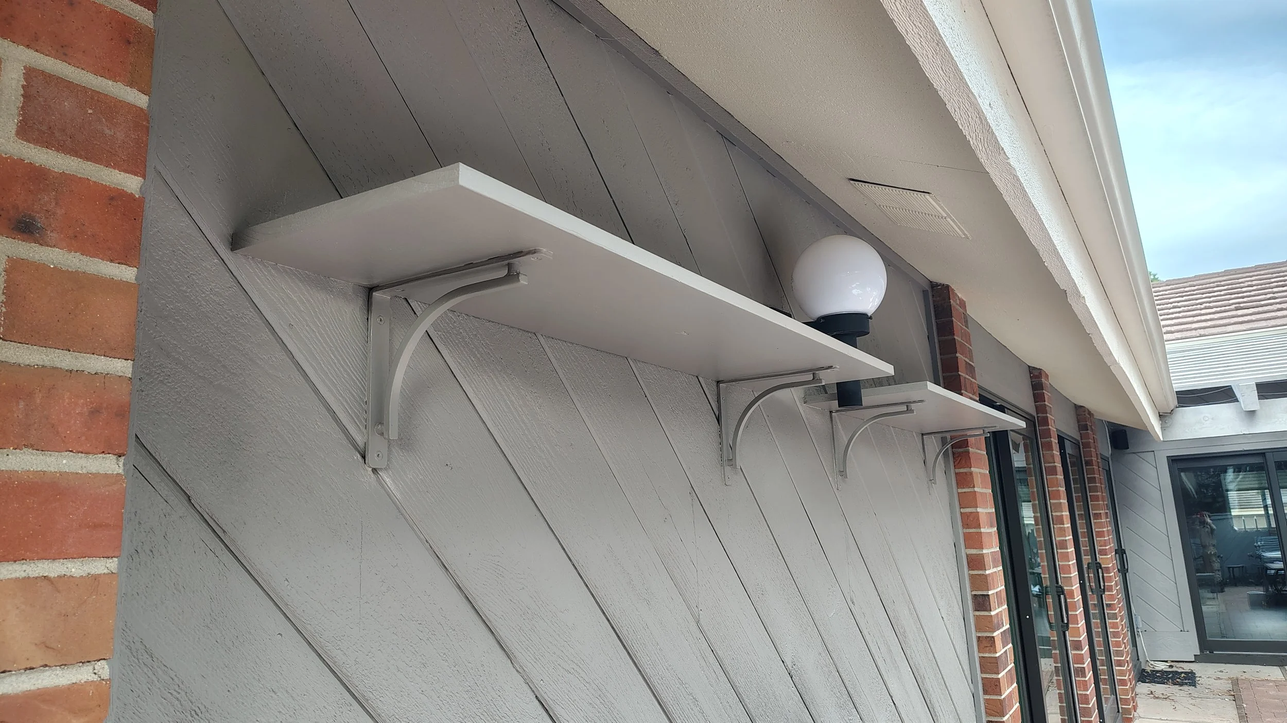 Outdoor house wall with three white wall-mounted shelves, brick accents, and a light fixture with a white globe. Glass sliding door with black framing is visible in the background.
