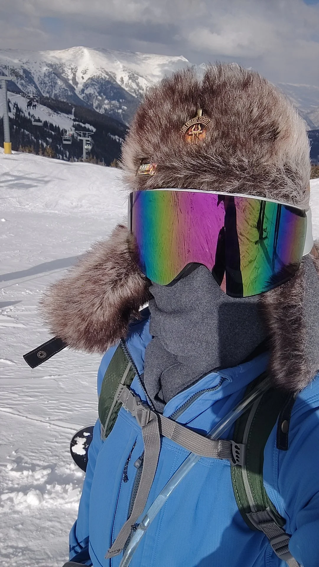 Person wearing a blue winter jacket, furry hat with decorative pins, and rainbow-tinted ski goggles in a snowy mountain landscape with ski lifts and snow-covered peaks.
