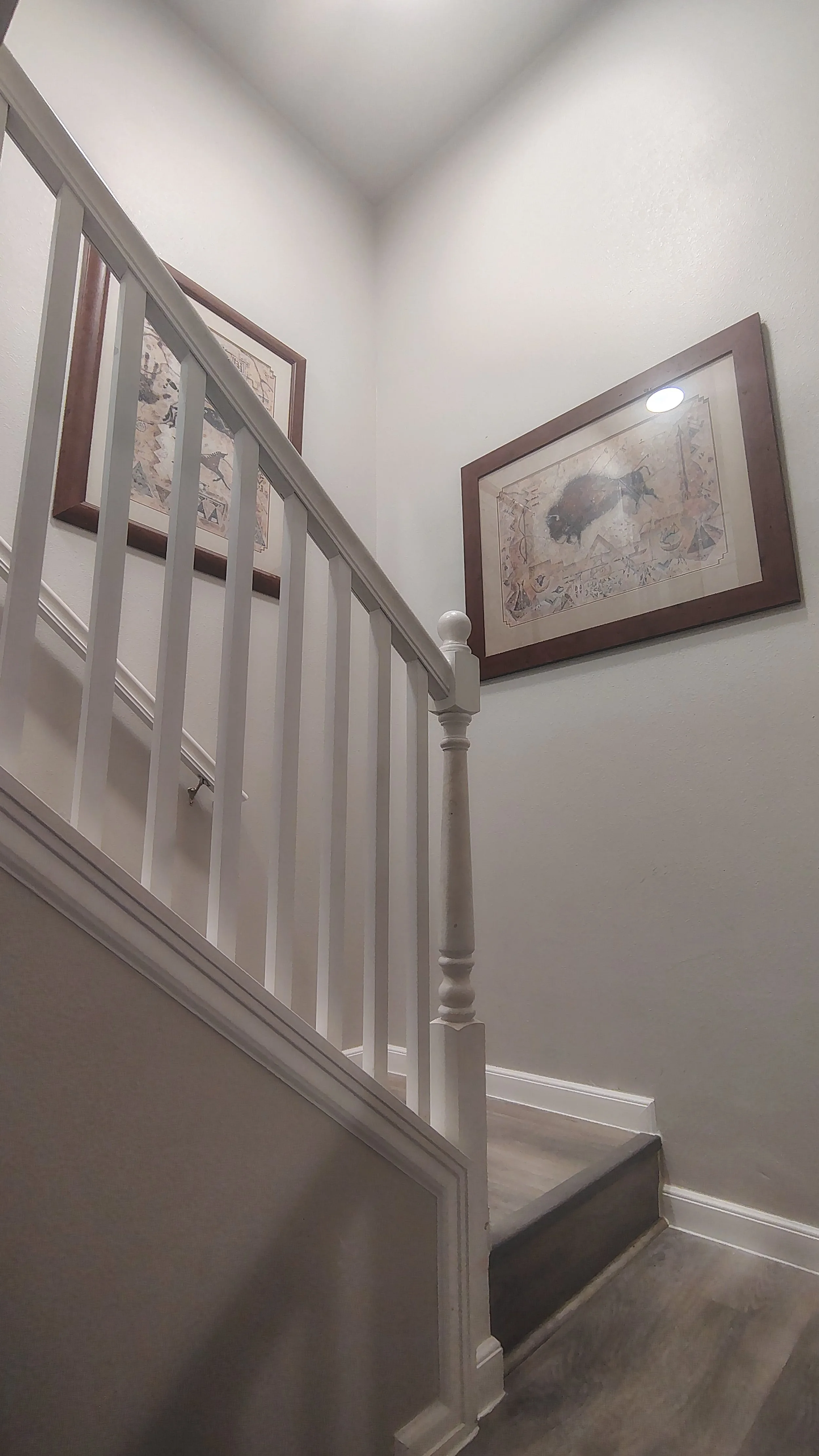 Interior view of a staircase with white walls, framed artwork on the wall, and a wooden floor.