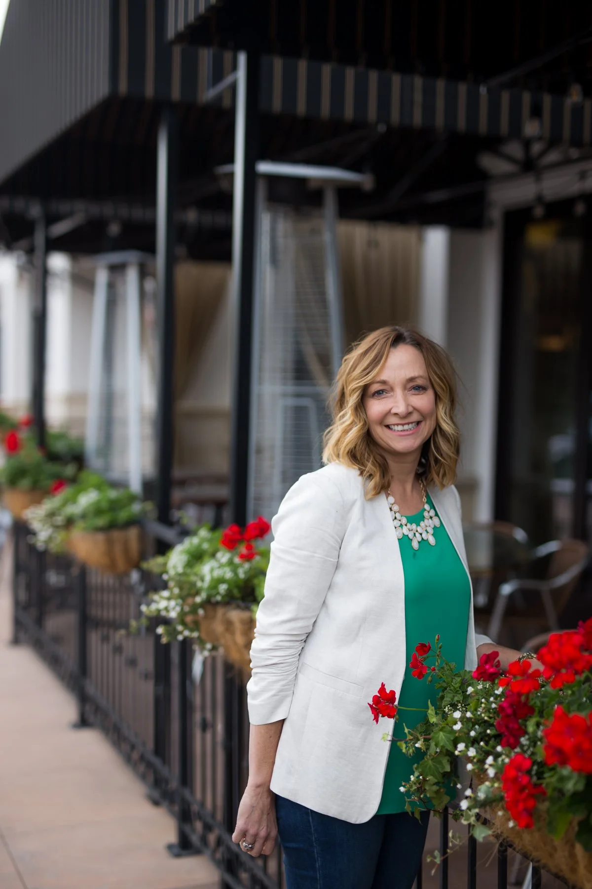 A woman with shoulder-length blonde hair smiling on a sidewalk near a restaurant or café with flower baskets, wearing a white blazer, green top, and a white necklace.