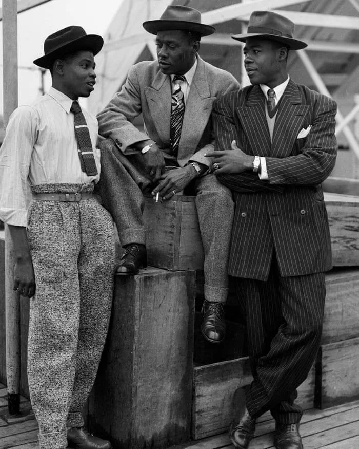 Three African American males, dressed in vintage clothing, standing and sitting outdoors on a wooden structure, engaged in conversation.
