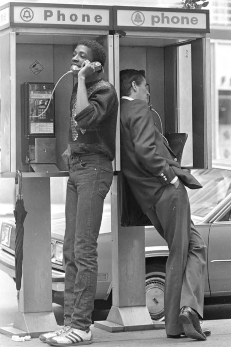 Two men at separate phone booths, one smiling and talking on a corded phone, the other leaning against the booth, looking away, with a phone in his hand.