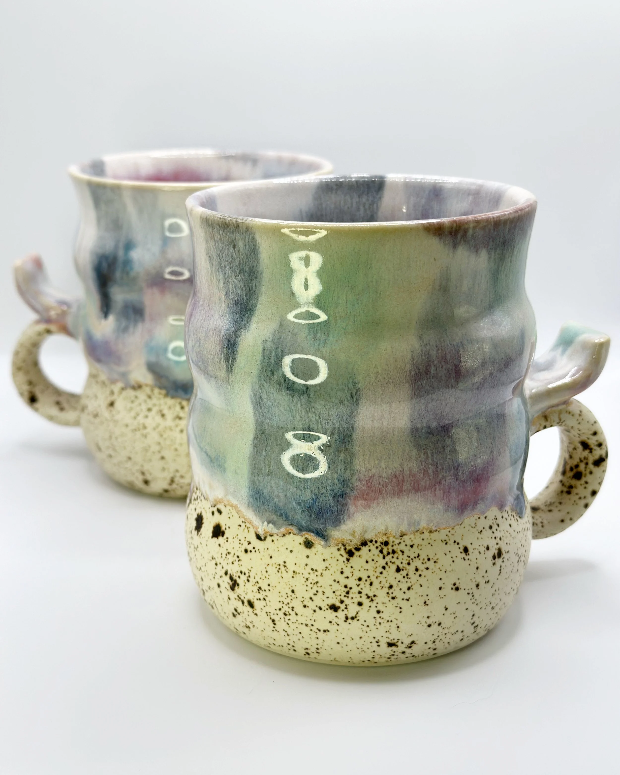 Two ceramic mugs with abstract watercolor-like glaze, each with a speckled beige lower half and a small handle, set against a white background.