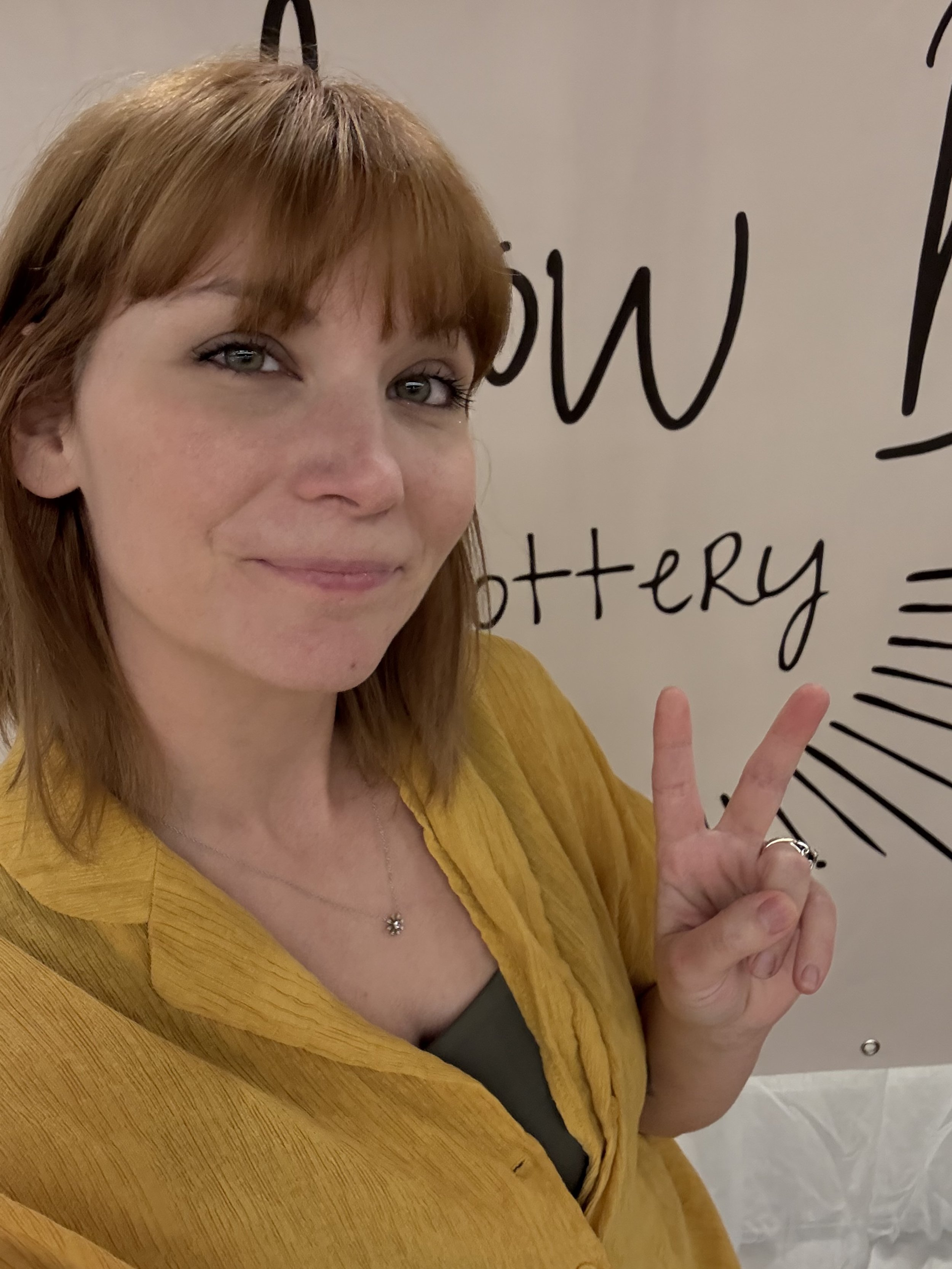 A woman with red hair and a yellow shirt making a peace sign gesture in front of a sign that says 'Show Pottery'.