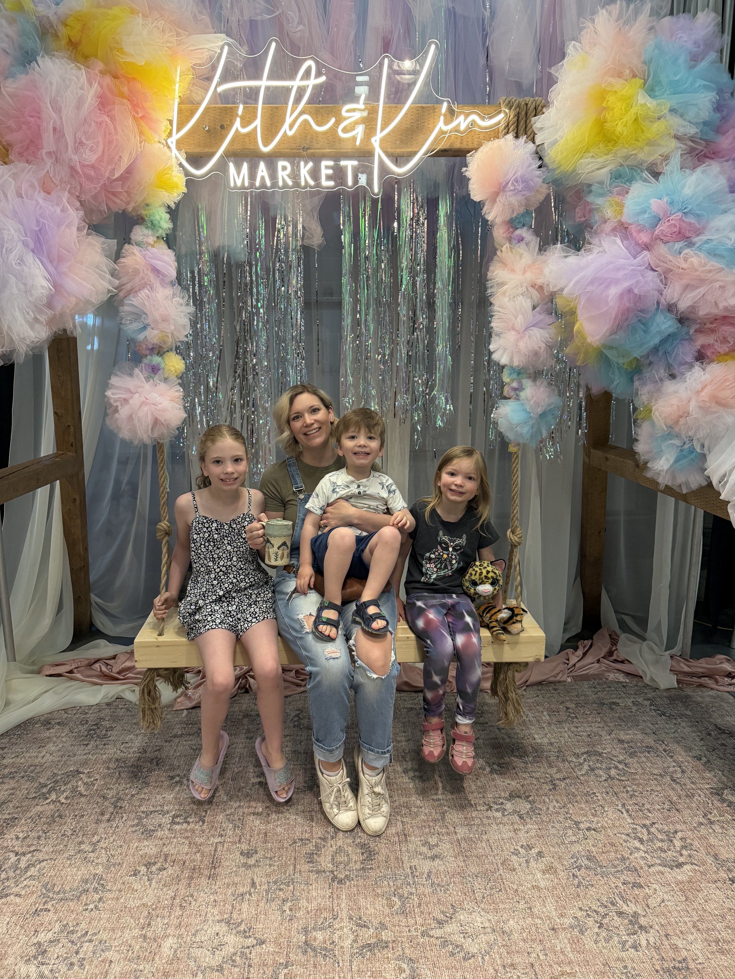 A woman with three children sitting on a wooden swing at a colorful, decorated indoor event, with a neon sign reading 'Lithevin MARKET' in the background and pastel-colored tulle and metallic streamers decorations.