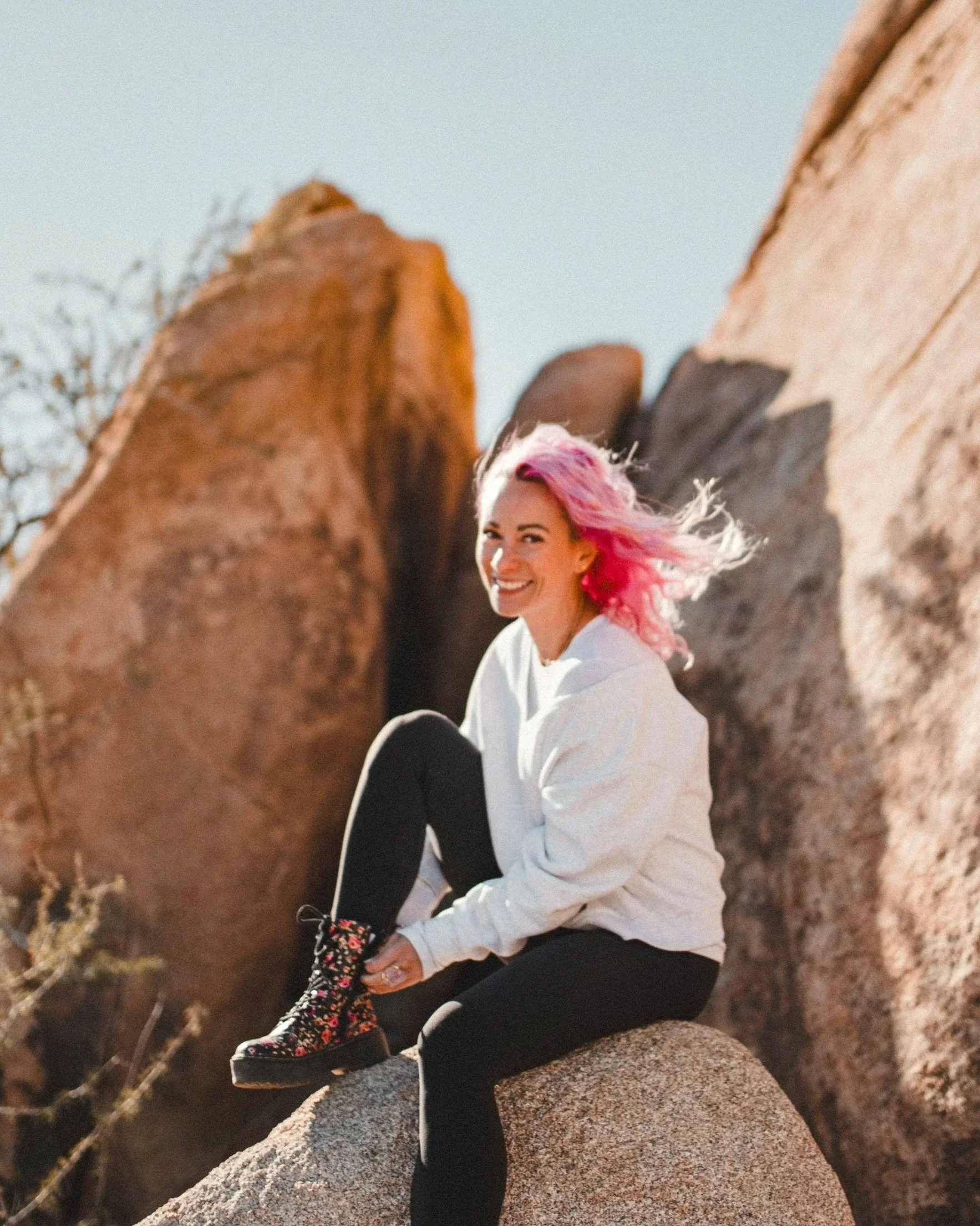 Woman with pink hair sitting on a rock in a desert landscape with large boulders and clear sky.