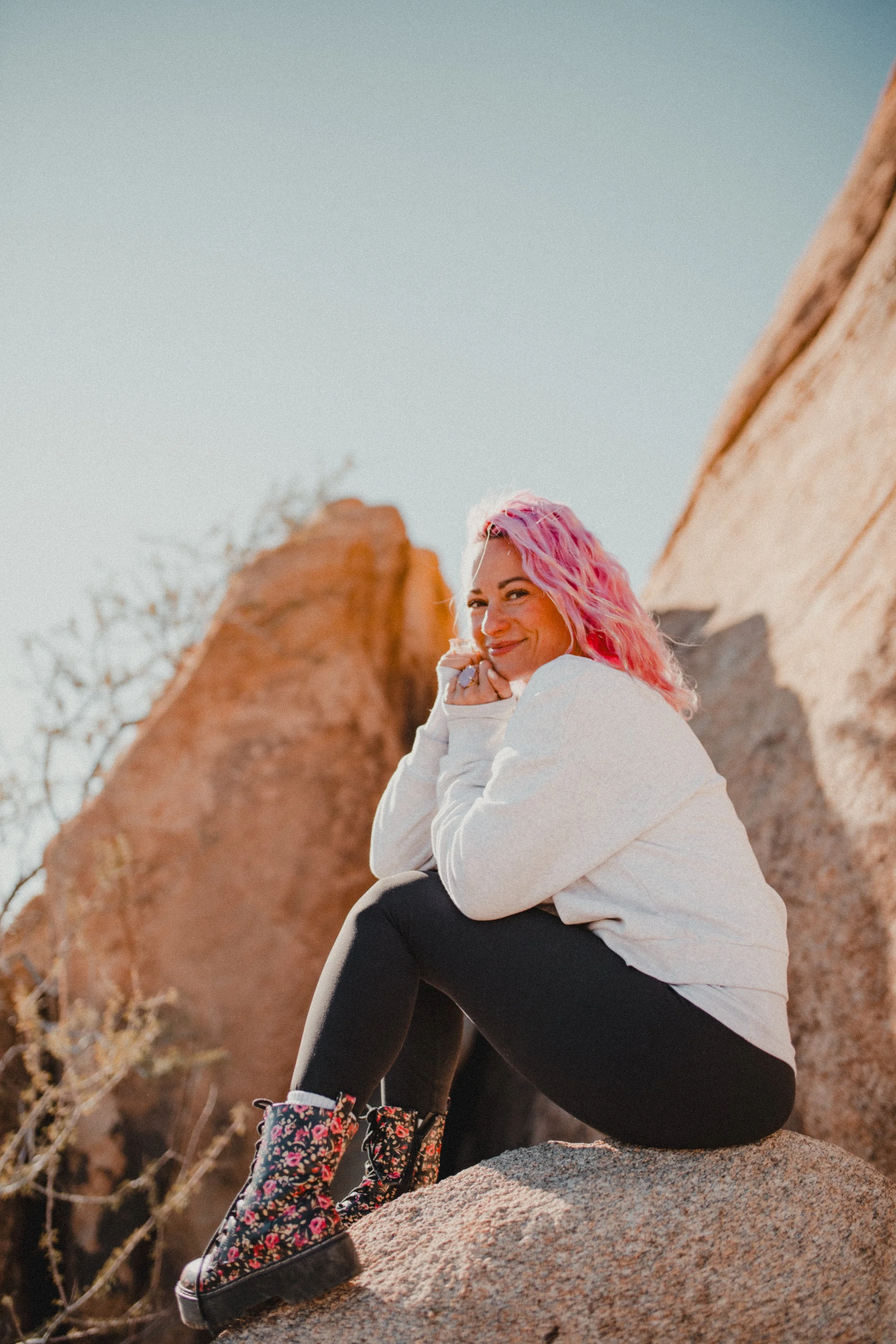A woman with pink hair sitting on a rock outdoors, surrounded by desert landscape and large rock formations, wearing a white hoodie, black leggings, and floral hiking boots.