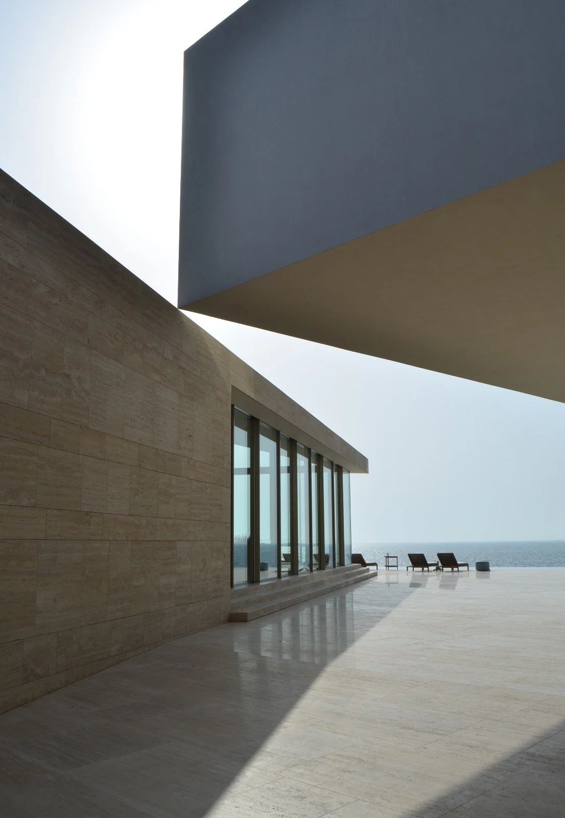 Modern building with large glass windows overlooking the ocean, with lounge chairs outside and a large shadow cast by an overhanging structure.