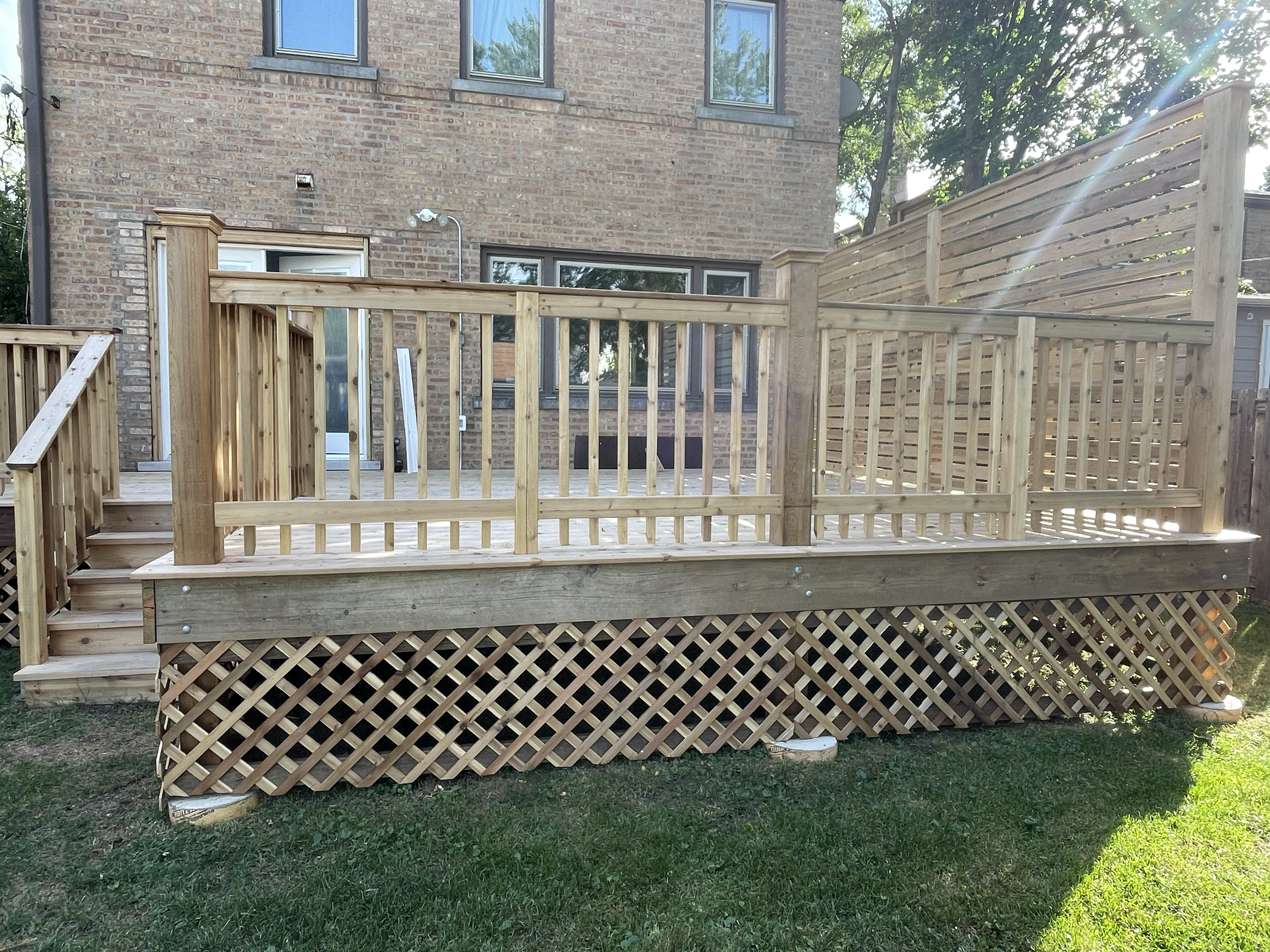New wooden deck with railing attached to the back of a brick house, with steps leading down to grass and a lattice skirting below the deck, in a backyard.