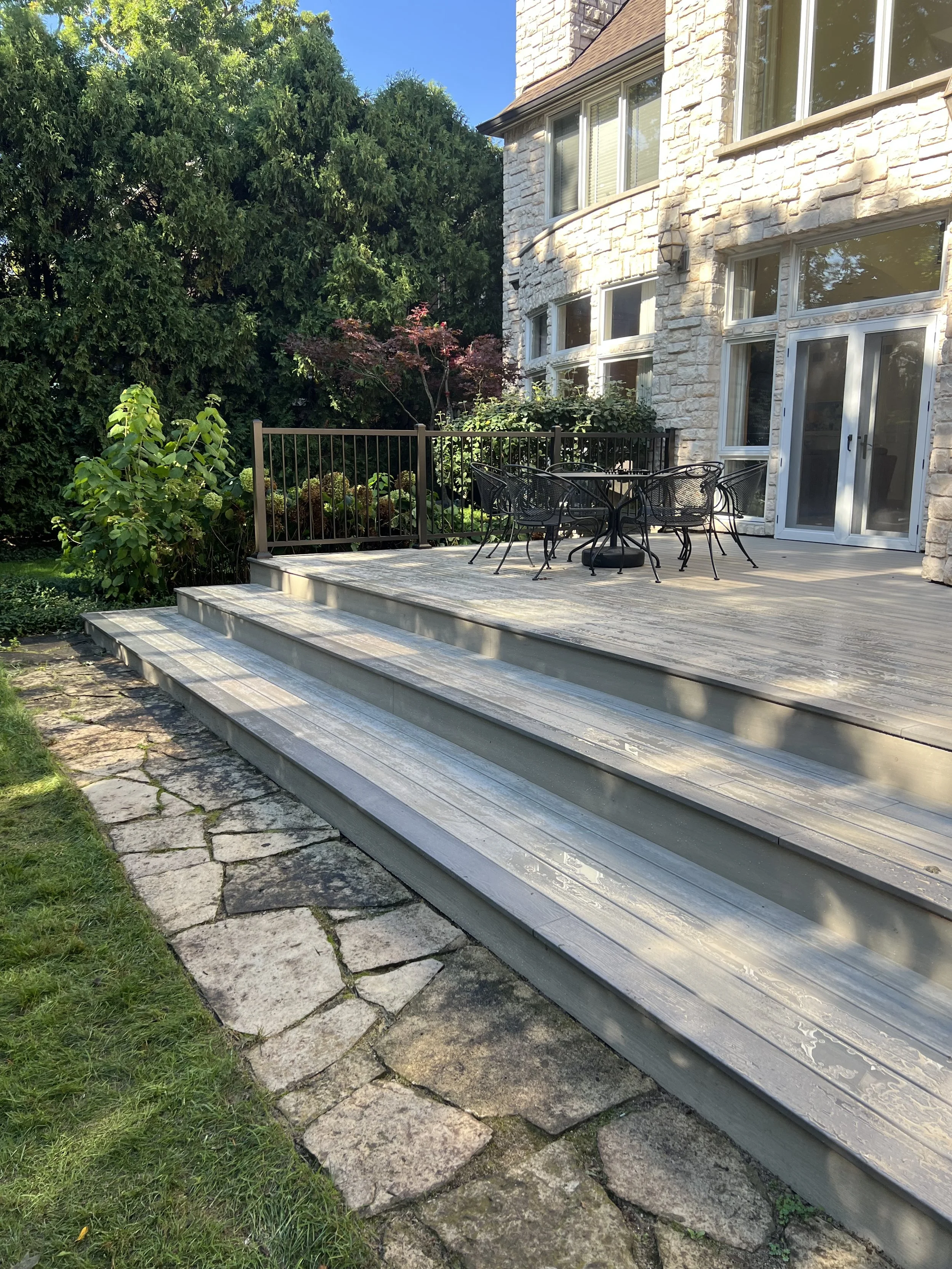 Backyard with a wooden deck, outdoor table and chairs, stone steps, lush greenery, and a stone house exterior.