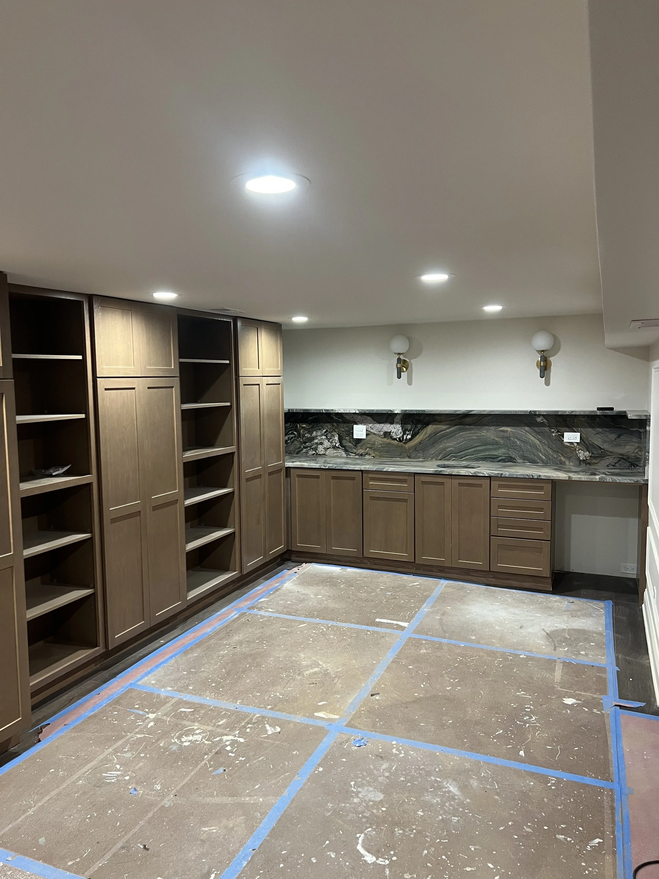 A room with wooden built-in cabinets and shelves, a marble backsplash, and a partially finished floor marked with blue tape, indicating ongoing renovation.