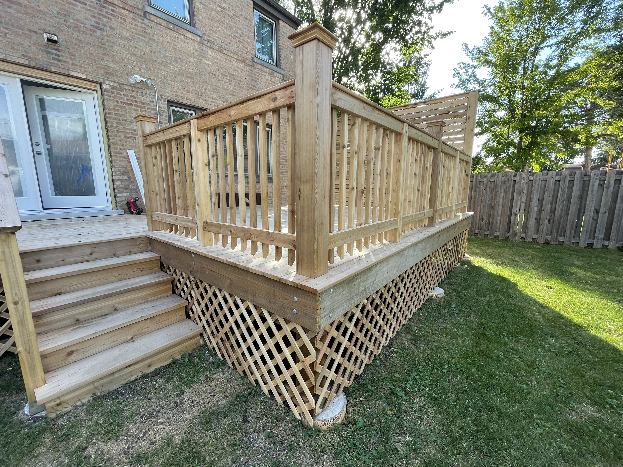 A newly built wooden deck with stairs leading to a backdoor of a brick house. The deck has a railing with vertical balusters and lattice skirting underneath, situated on grass with a wooden fence in the background. Trees are visible behind the fence.