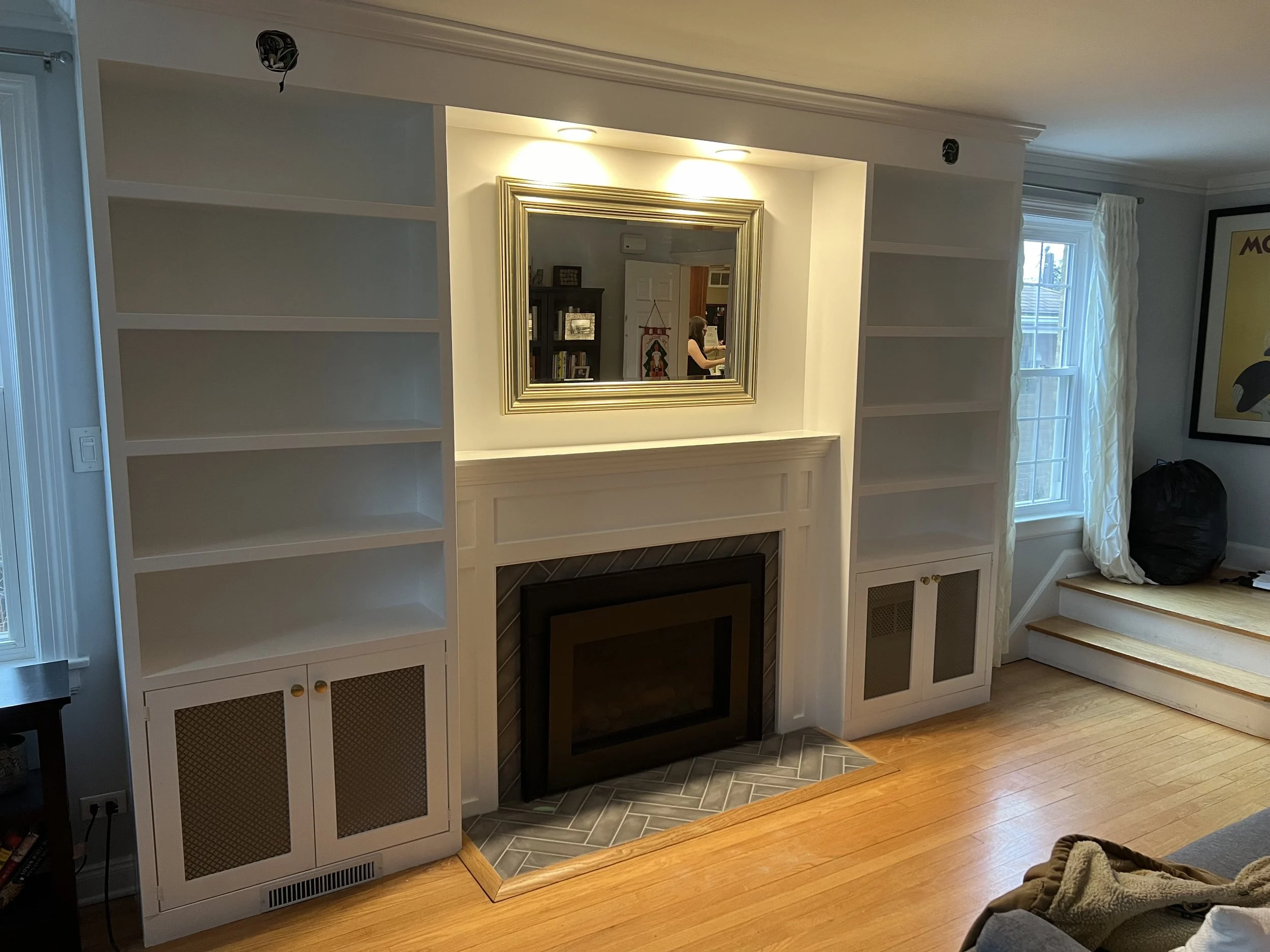 Living room with built-in white bookshelves on both sides of a fireplace, a mirror above the fireplace, and a window with curtains to the right.