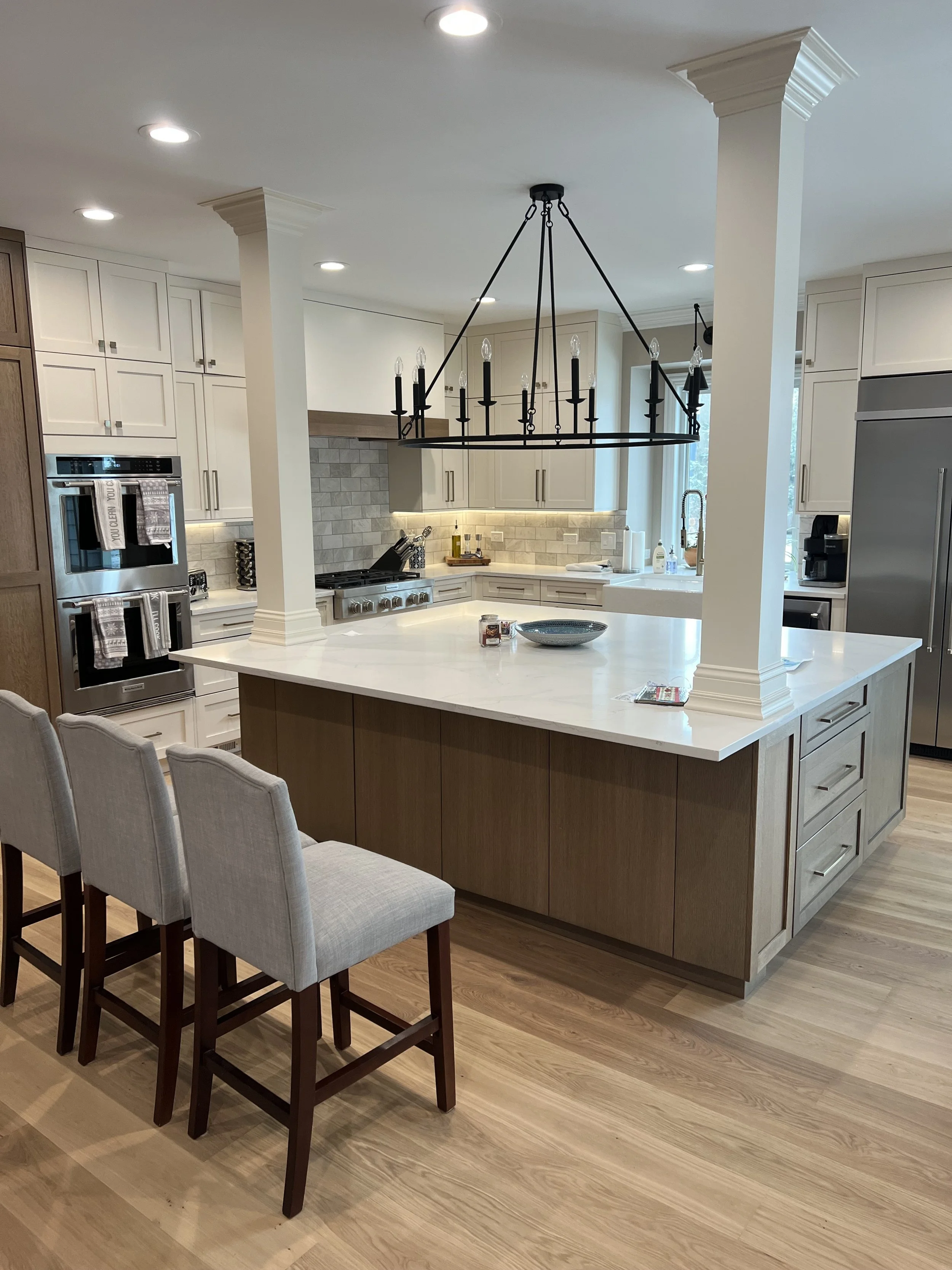 Modern kitchen with island, bar stools, chandelier, stainless steel appliances, off-white cabinets, tile backsplash, and wooden flooring.