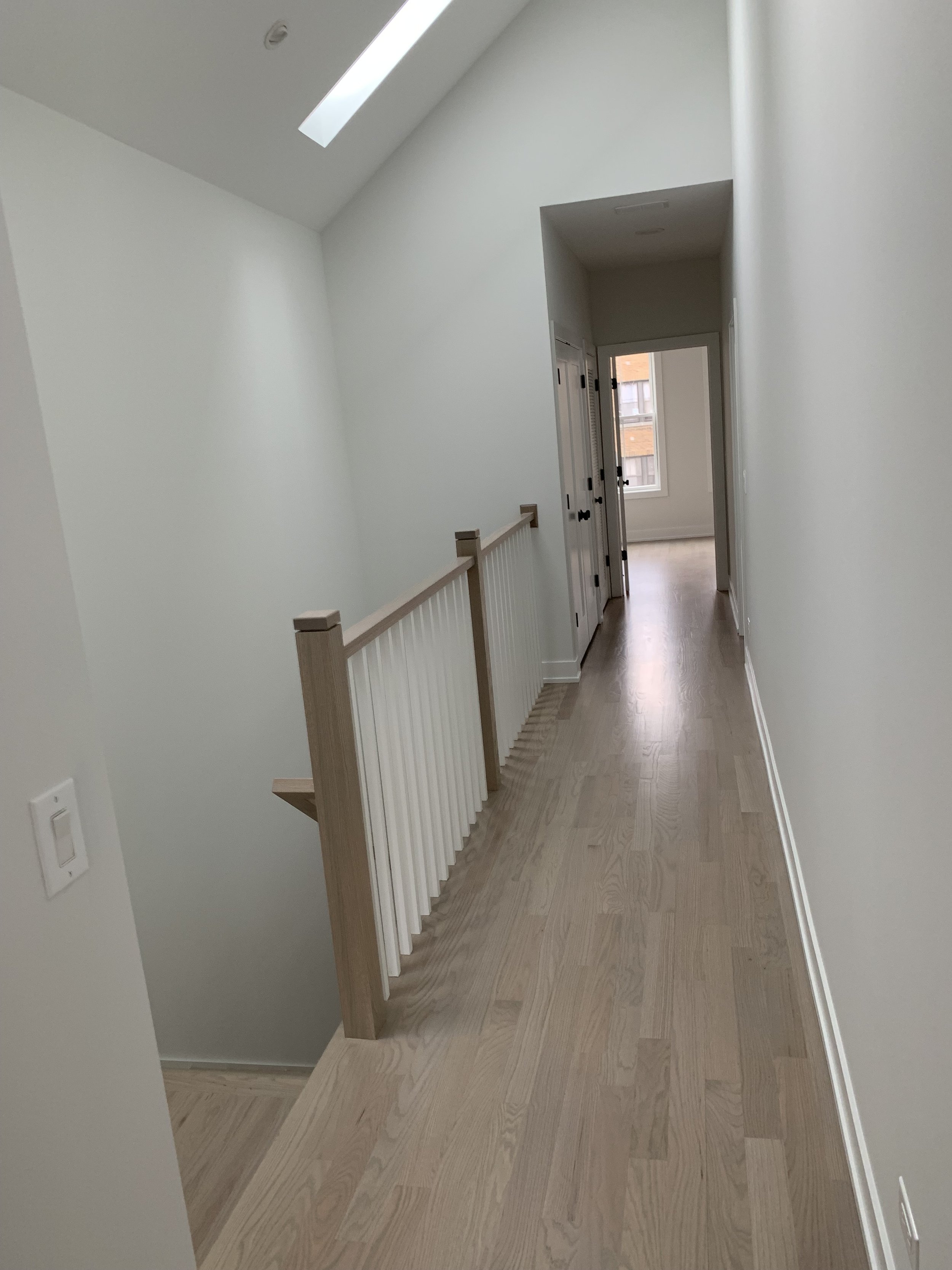 Bright hallway with wooden flooring, white painted walls, skylight, and a staircase with a white railing on the left.