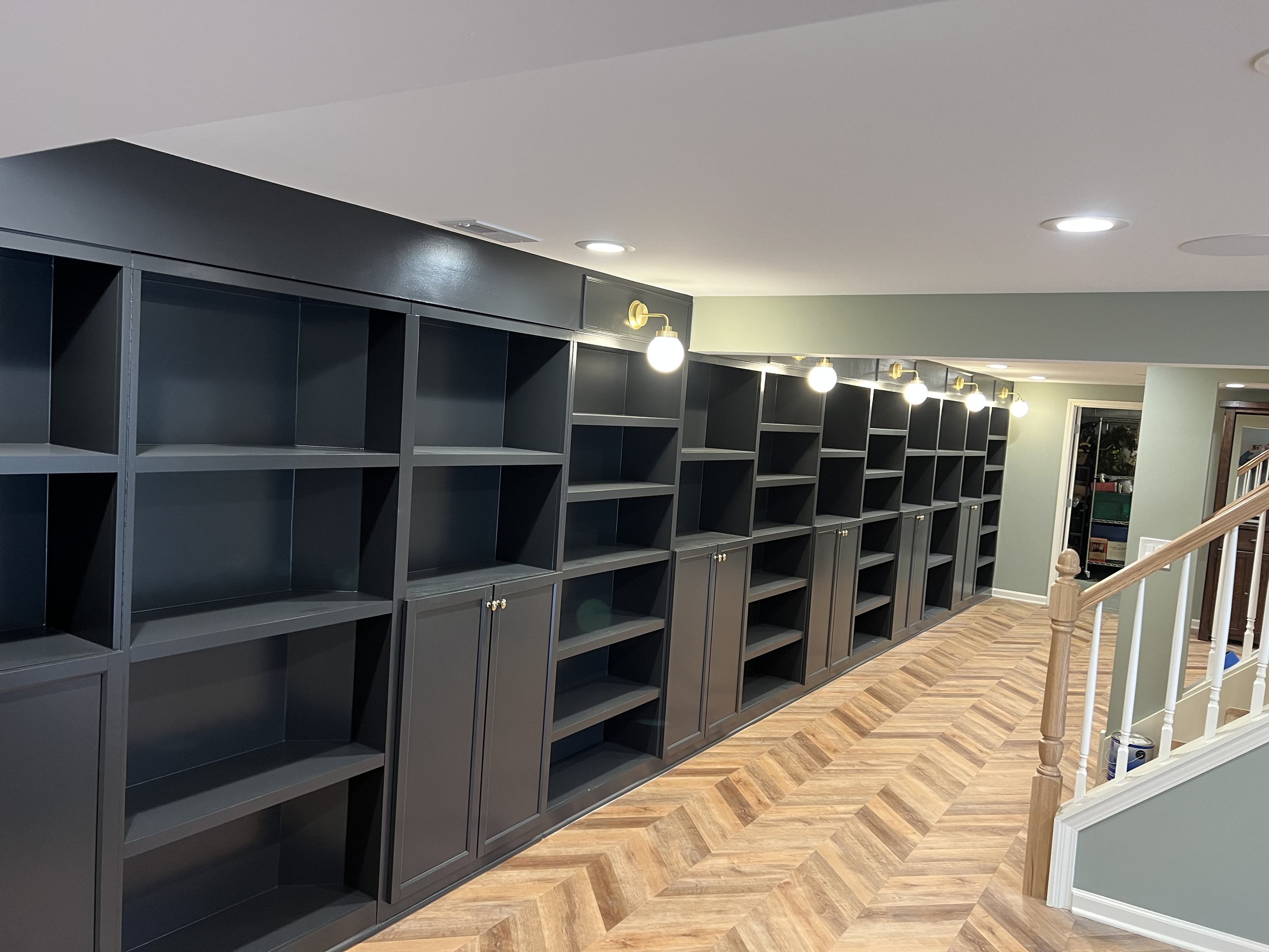 A long row of empty black bookshelves with closed cabinets at the bottom, located in a well-lit room with wooden floor and a staircase to the right.