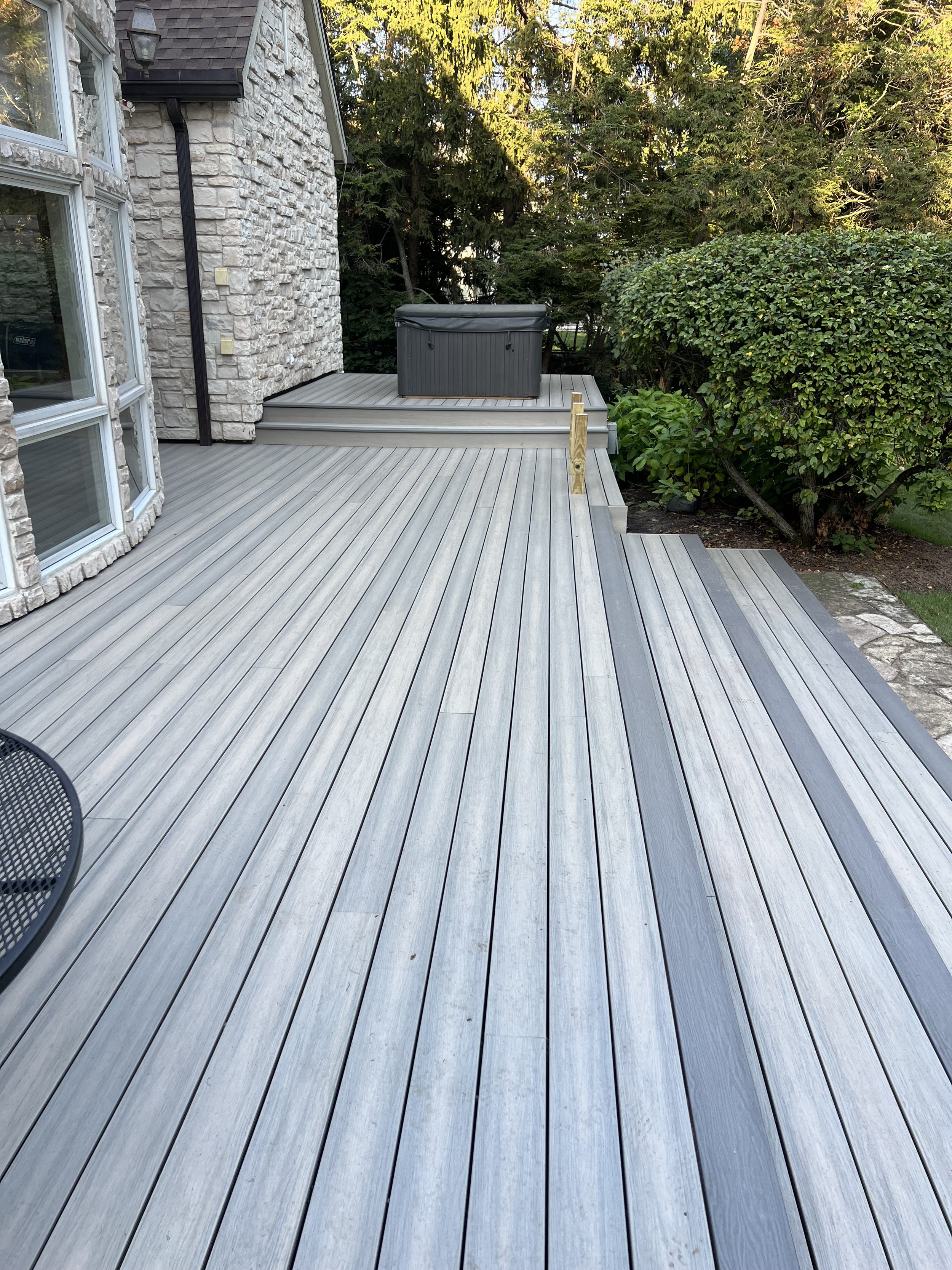 A wooden deck attached to a house with a stone exterior wall, a hot tub in the background, and lush green bushes and trees surrounding the area.