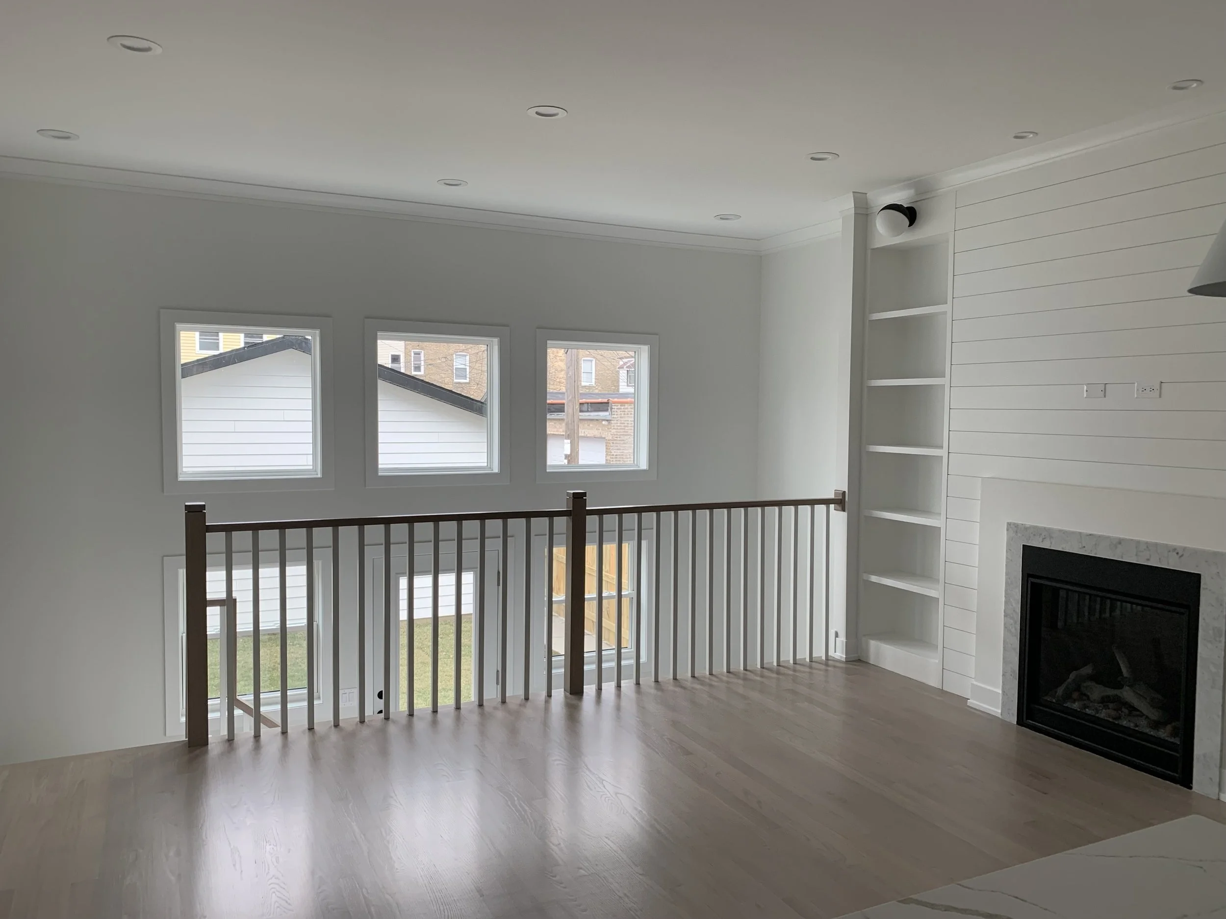 Empty living room with three windows, white walls, a fireplace with a white surround, built-in white shelves, wooden flooring, and a railing overlooking a lower level.