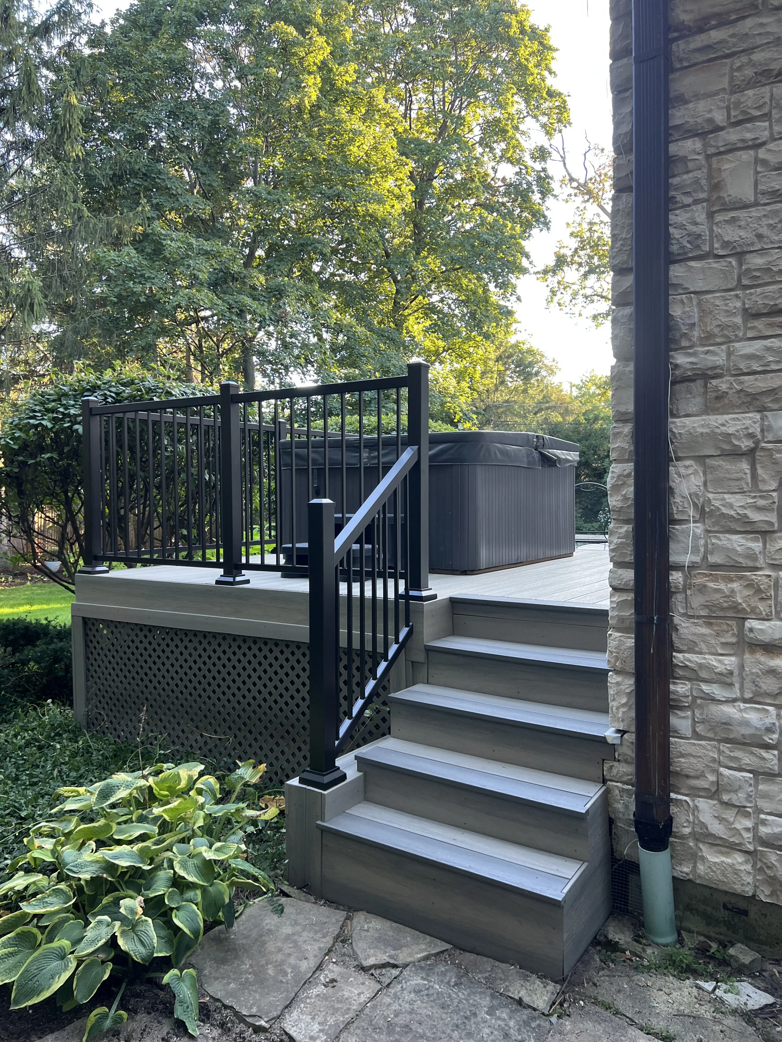A small wooden staircase with black metal railings leading to a deck with a hot tub, next to a stone house wall, with greenery and trees in the background.