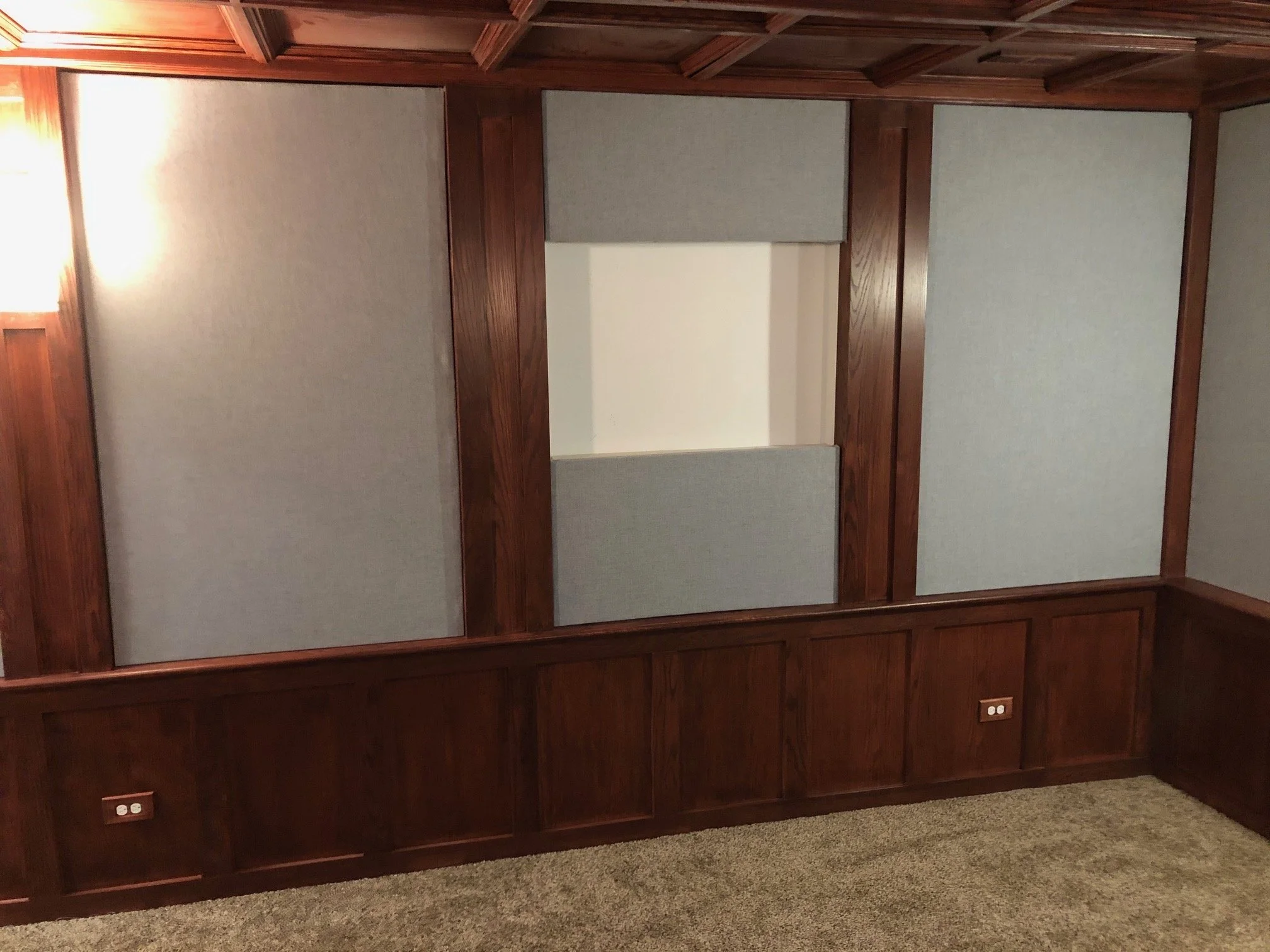 Empty room with wood-paneled walls, gray acoustic panels, carpeted floor, and two electrical outlets.